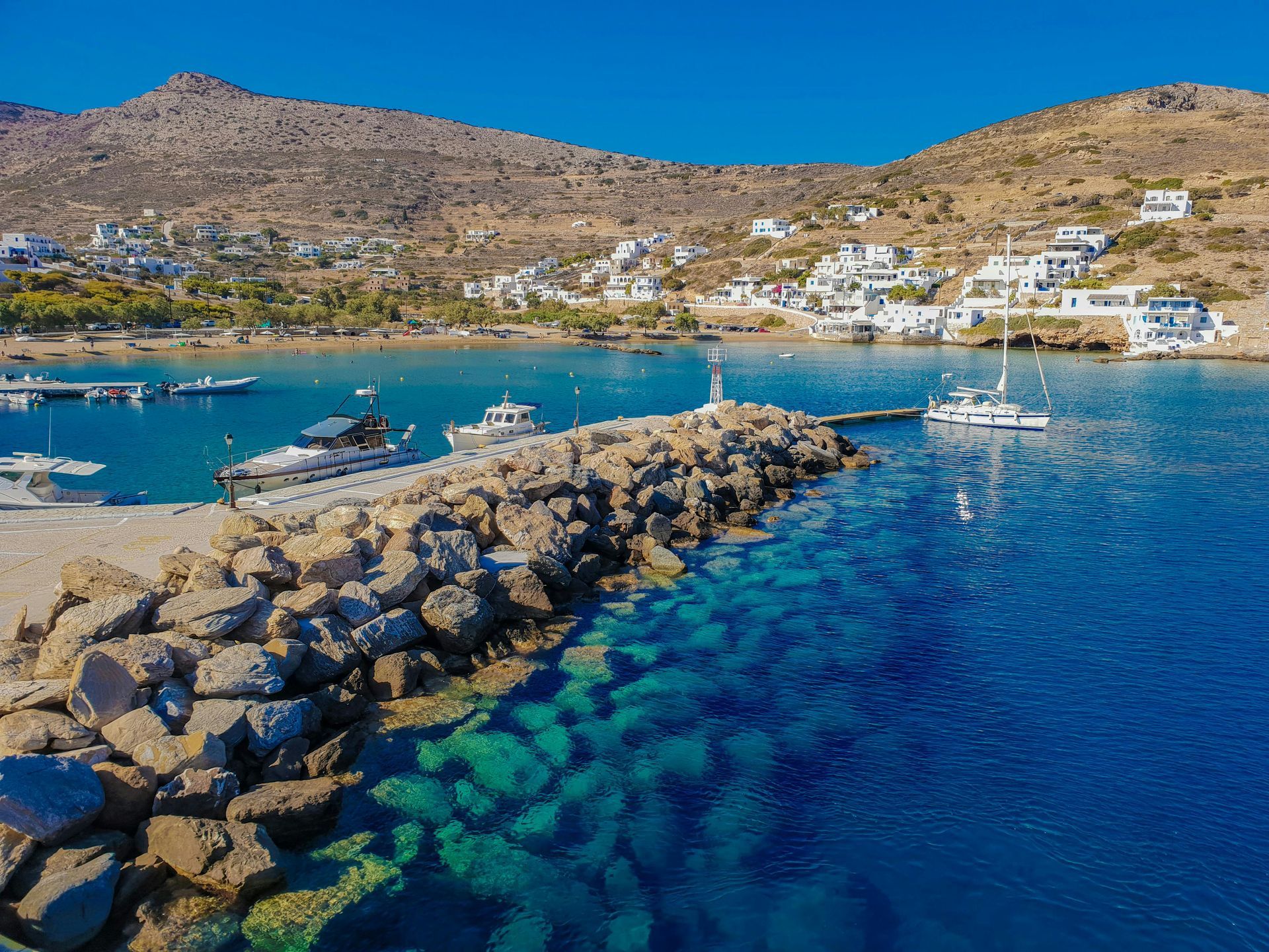 Coastal town with boats docked at a rocky pier, clear turquoise water, and mountains under a blue sky.