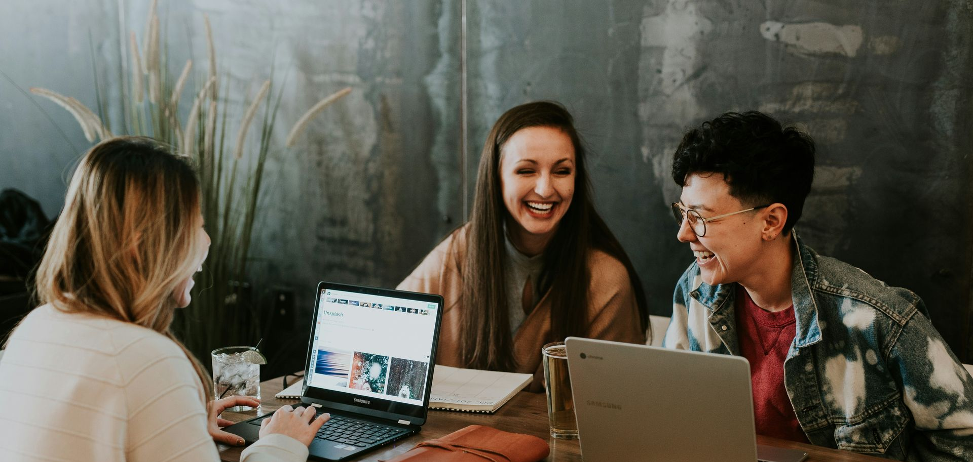 Three women laughing around a table, working on laptops. The setting appears to be a cafe.