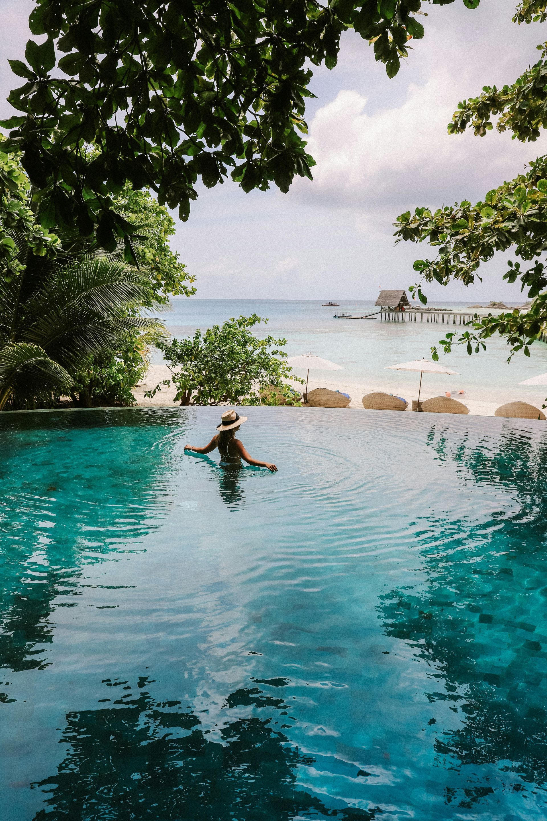 Woman in a pool with ocean view; turquoise water, white sand beach, trees frame the scene.