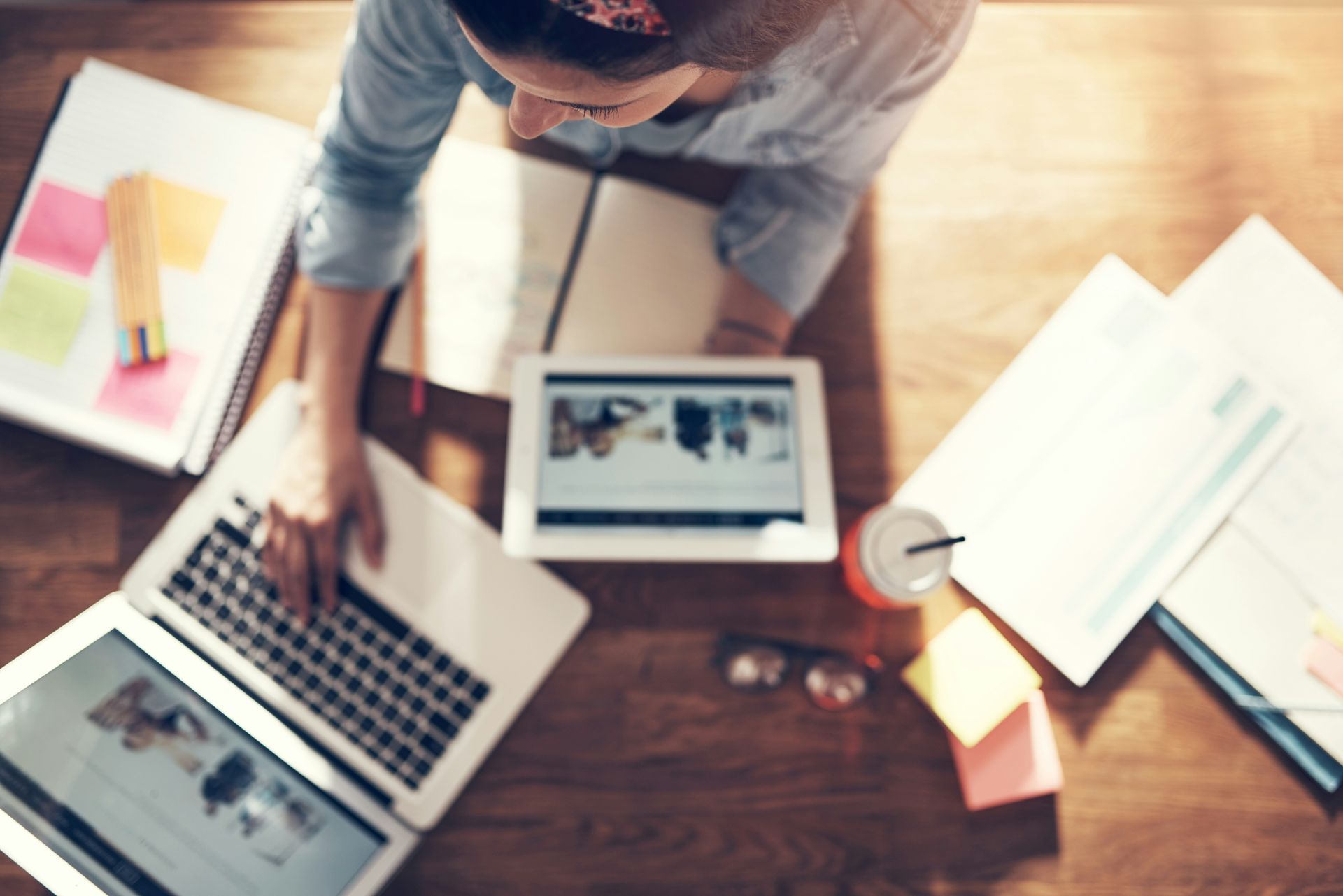 Person at a desk, using laptop and tablet, surrounded by papers, sticky notes, and a drink.