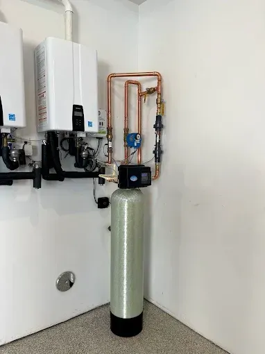 Wall-mounted water heaters and copper pipes above a tall cylindrical water softener in a utility room