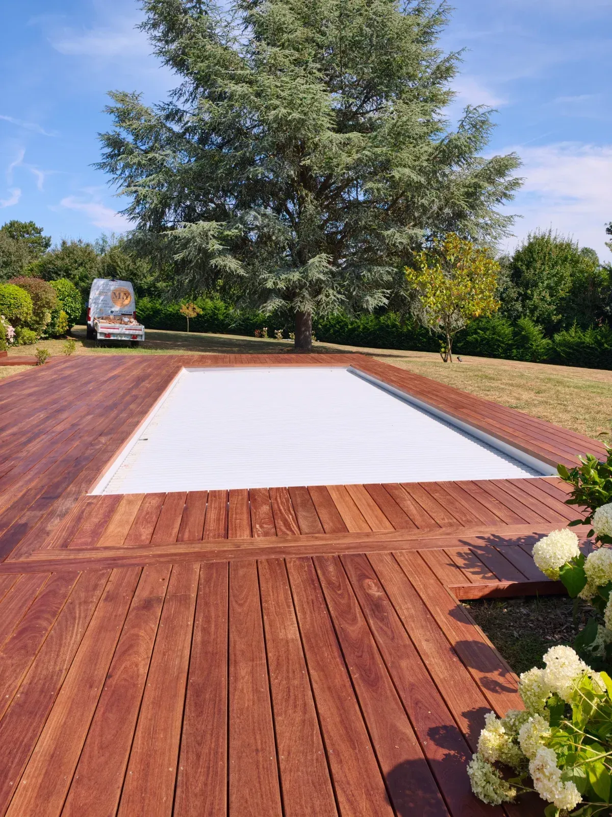 Plage de piscine en bois exotique avec volet roulant, camionnette MY Menuiserie en arrière-plan