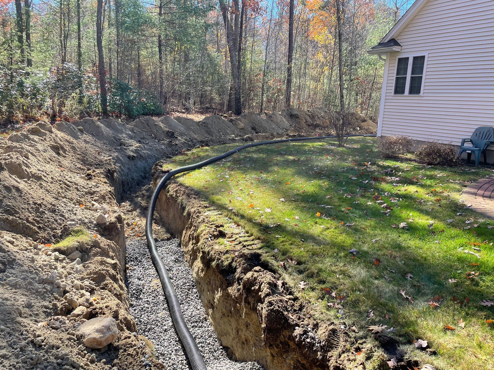 A black pipe in a trench with gravel, next to grass and a house, outdoors in daylight.