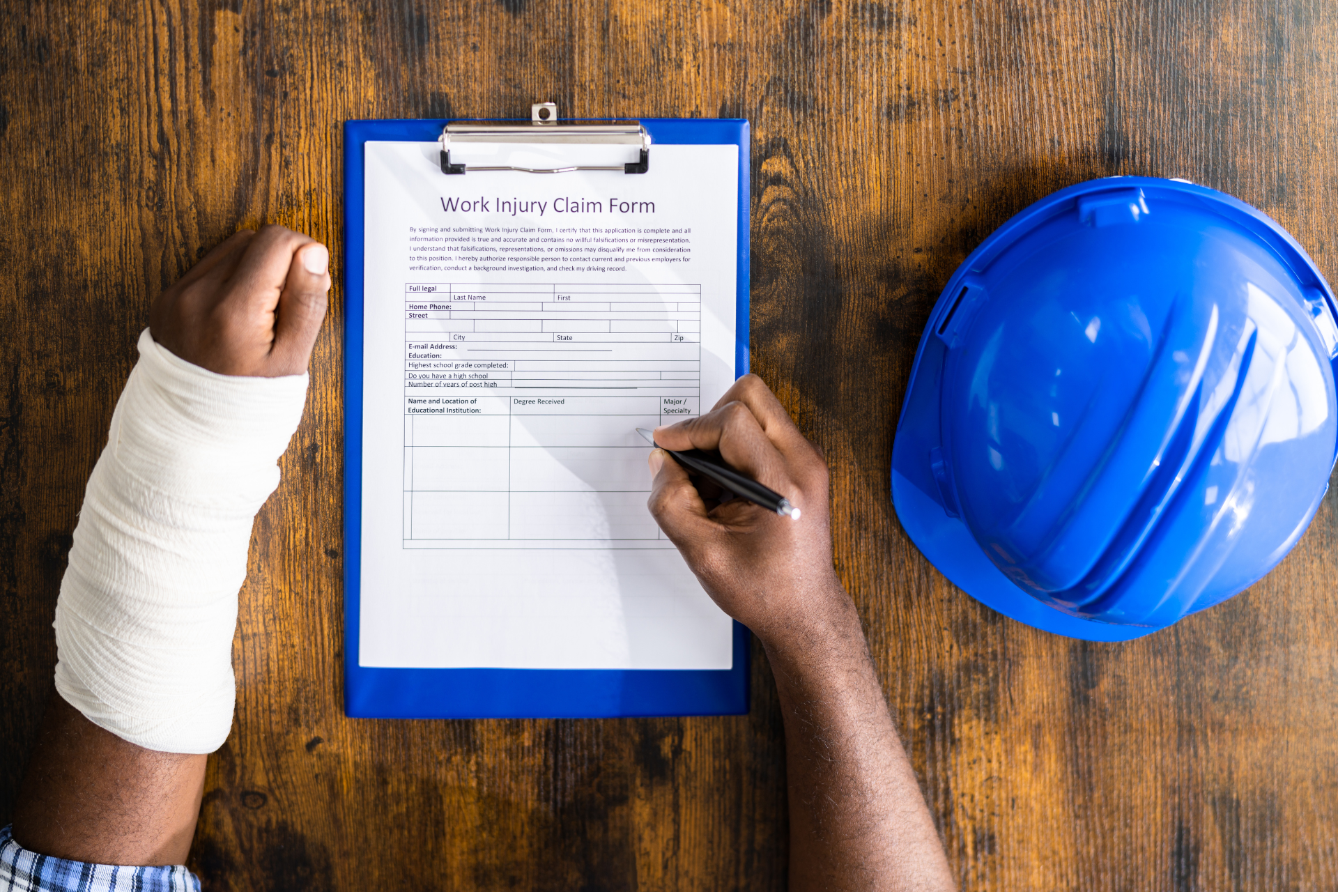A man with a cast on his arm is writing on a clipboard next to a hard hat.