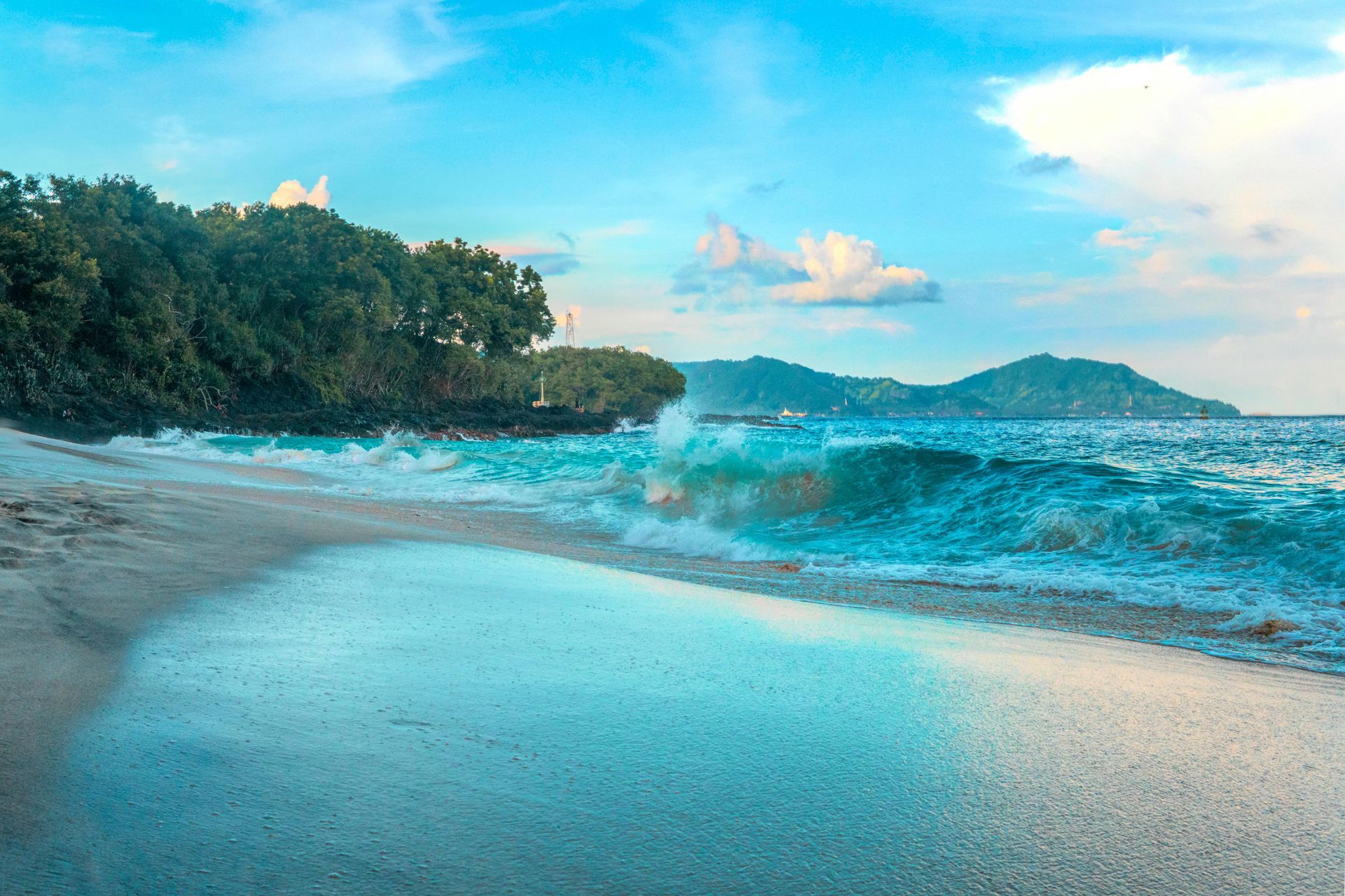 Beach scene: turquoise waves crash on white sand, under a blue sky, with trees on the left and mountains in the distance.
