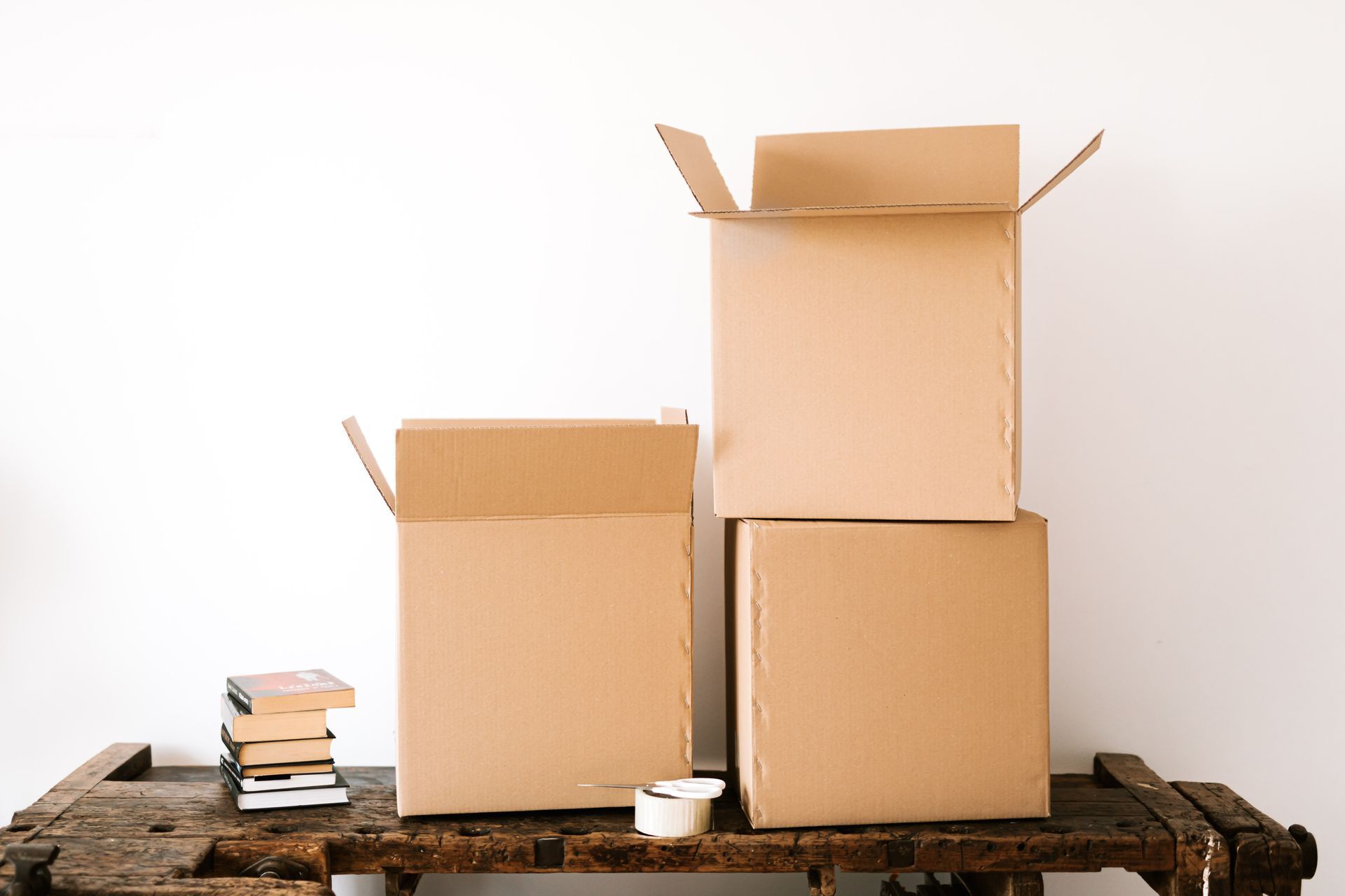 Tres cajas de cartón apiladas y una pequeña pila de libros sobre una mesa de madera contra una pared blanca.