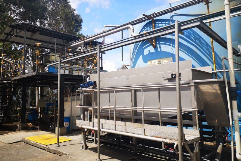 Planta de tratamiento de agua industrial con tanques, tuberías y estructuras de soporte de acero inoxidable. Mural azul y blanco al fondo.
