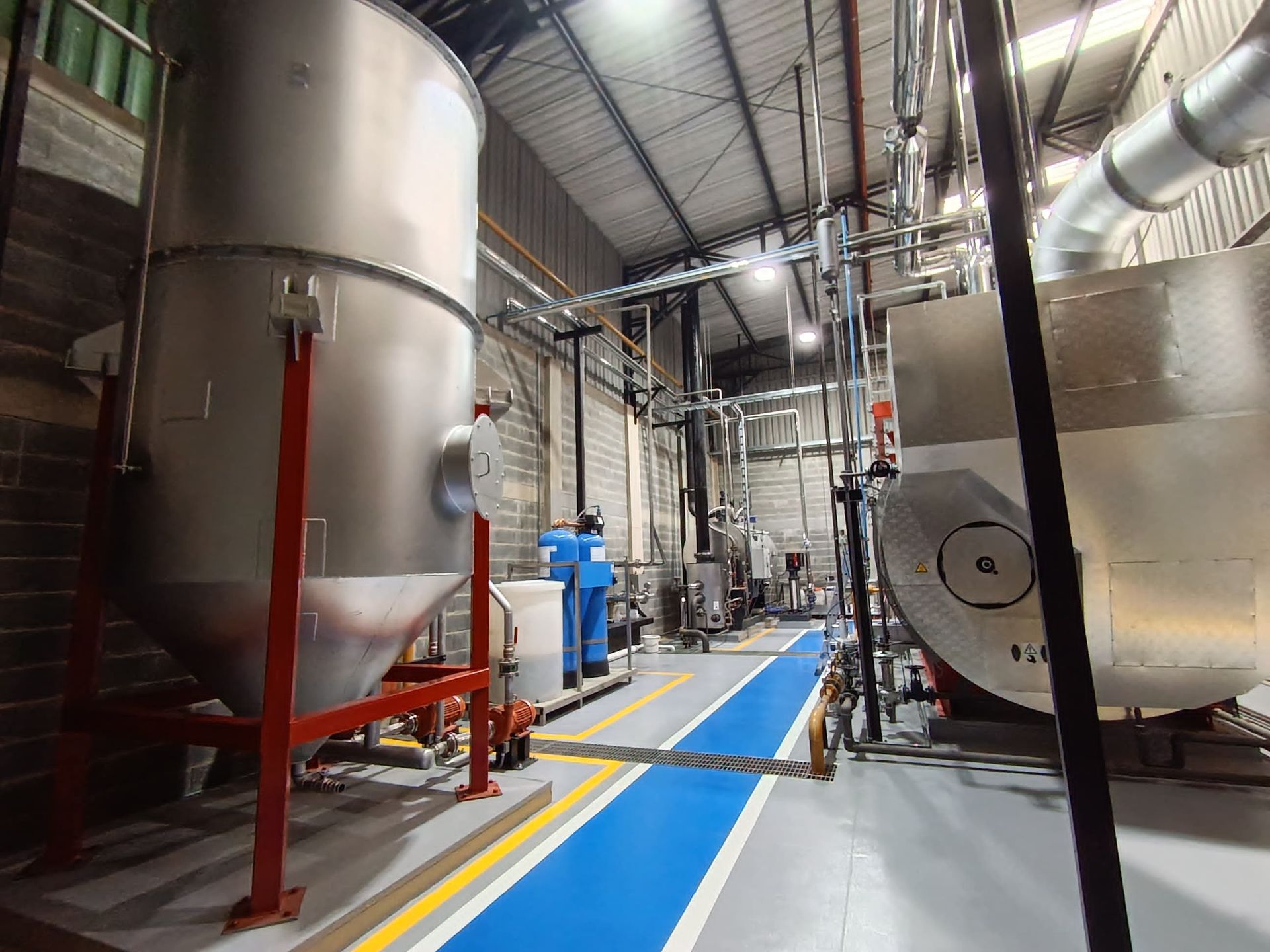 Interior de una instalación industrial con grandes tanques de metal, tuberías y camino azul; trabajador con uniforme azul.