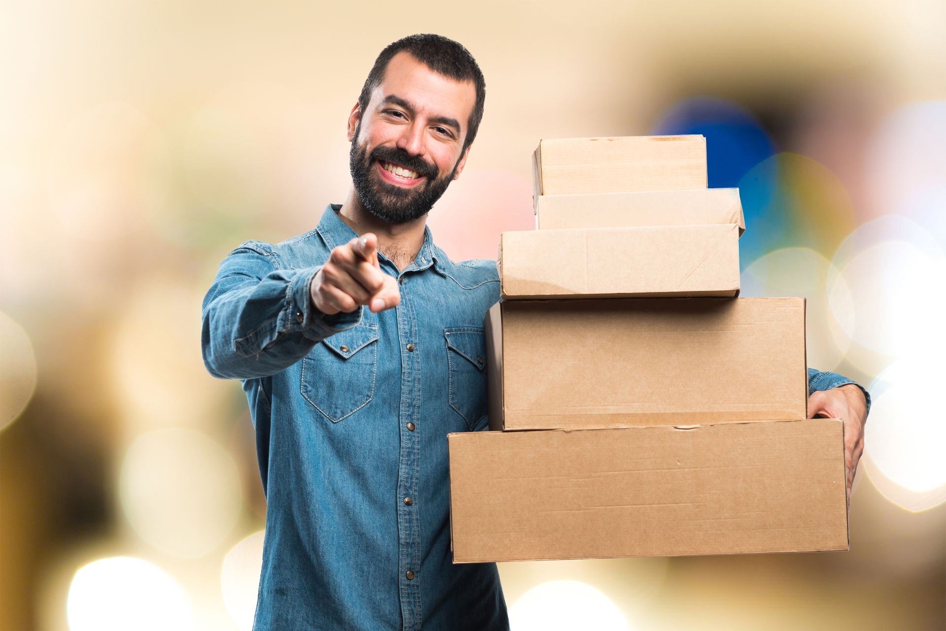 Hombre con camisa vaquera sosteniendo cajas de cartón apiladas, sonriendo y señalando al espectador. Fondo borroso.