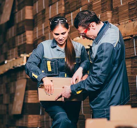 Dos trabajadores con uniforme de almacén abren una caja de cartón cerca de pilas de cajas.
