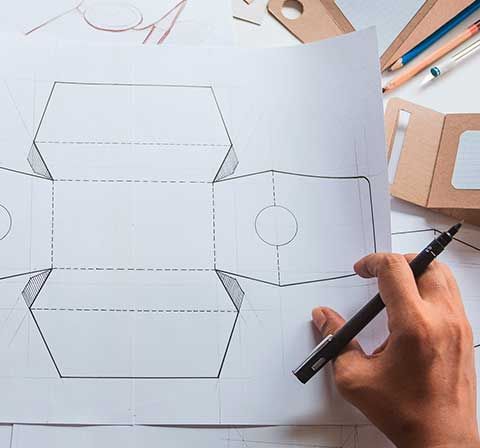 Dibujo a mano de una plantilla de caja de papel con un marcador negro sobre una superficie blanca, con otros elementos de diseño cerca.