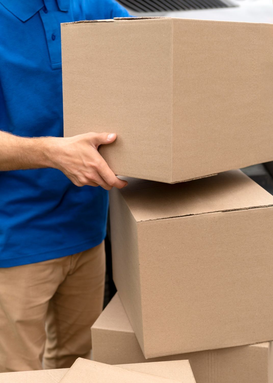 Un repartidor con camisa azul y pantalones color canela apila cajas de cartón cerca de un vehículo.