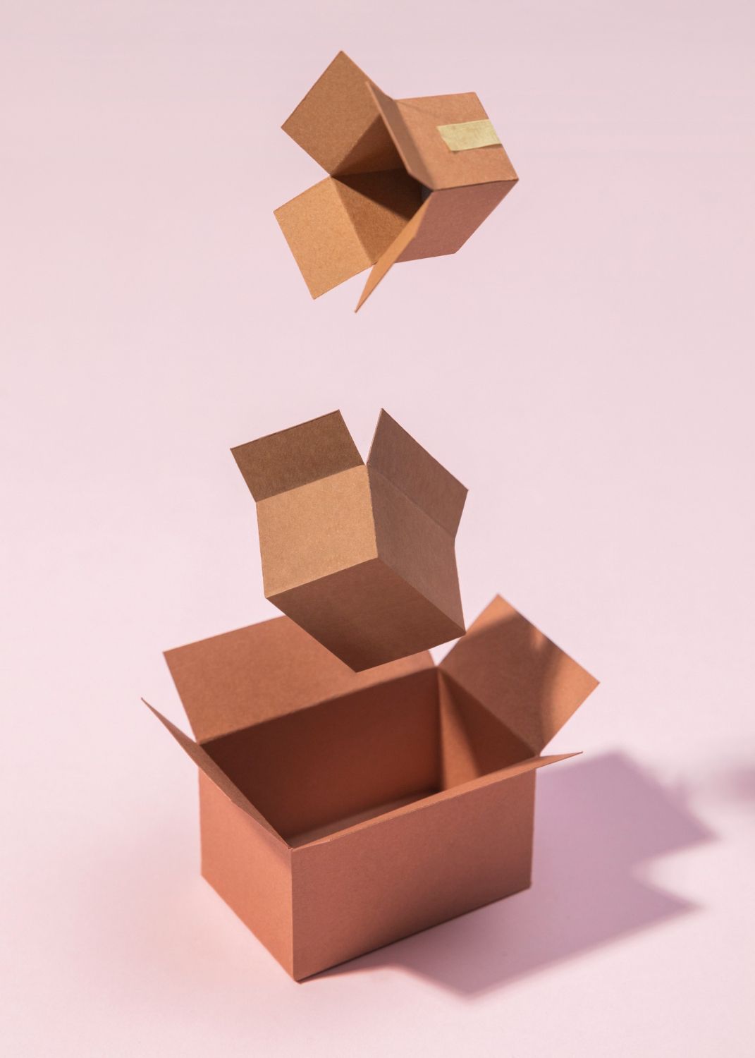 Tres cajas de cartón marrón, una abierta, una en el aire y otra encima con tapa pegada con cinta adhesiva, sobre un fondo rosa.