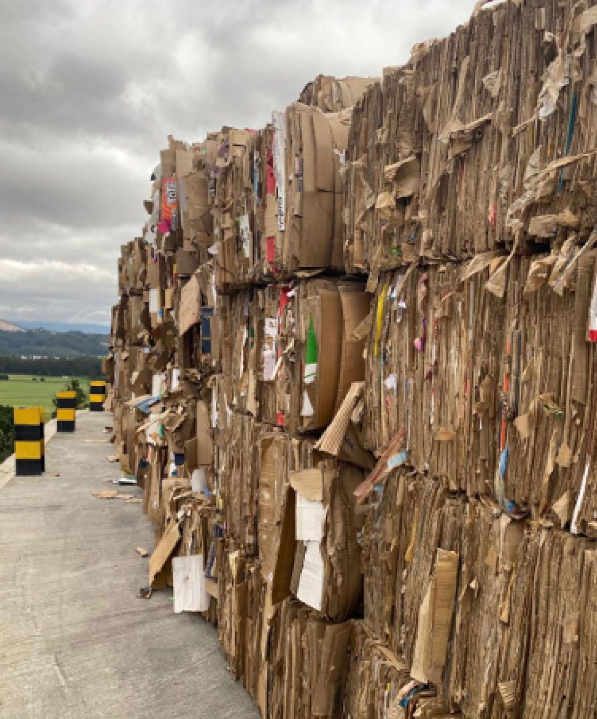Una gran pila de fardos de cartón comprimido en una instalación de reciclaje, con campos y un cielo nublado al fondo.