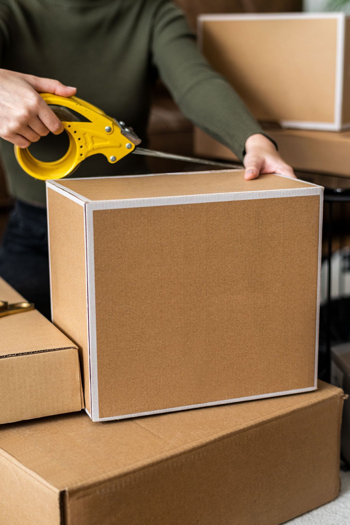 Persona colocando cinta adhesiva en una caja de cartón con una pistola de cinta amarilla. Las cajas están apiladas en un almacén.