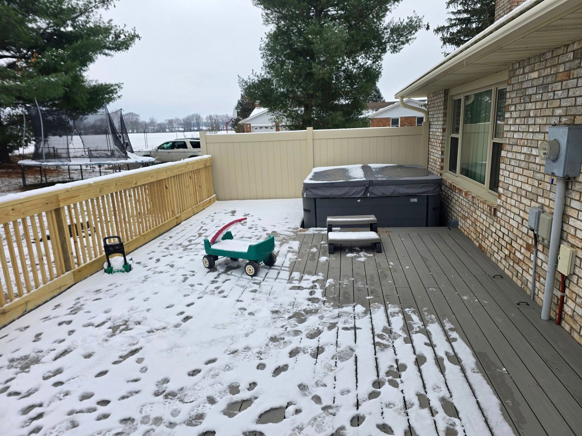 A snowy wooden deck featuring a hot tub, a child's green wagon, and a trampoline in the background.