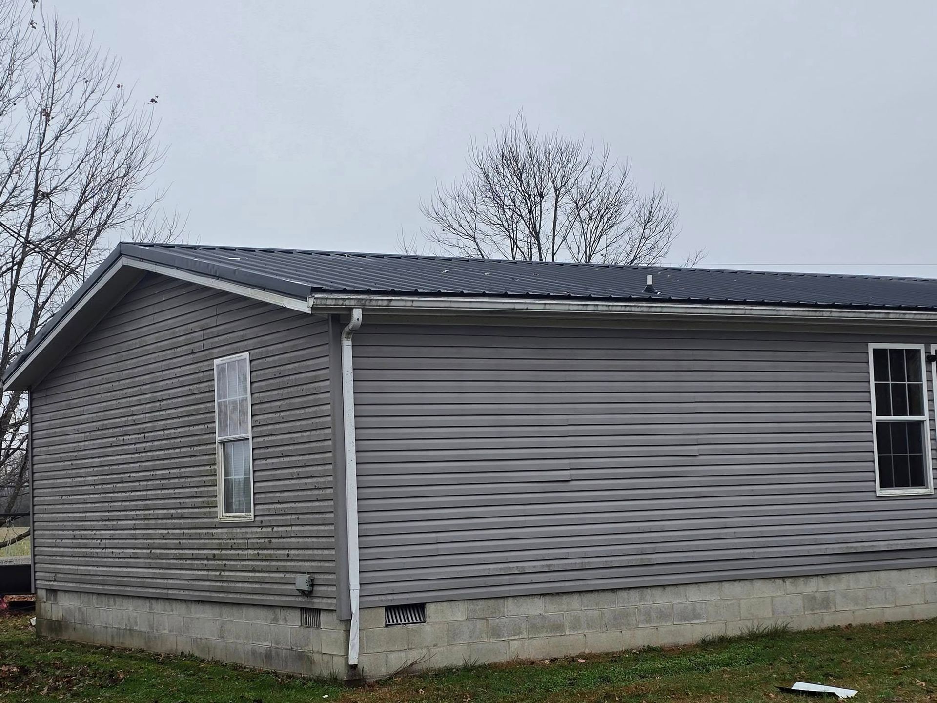 A side view of a single-story house with gray horizontal siding, a dark metal roof, and a concrete block foundation.