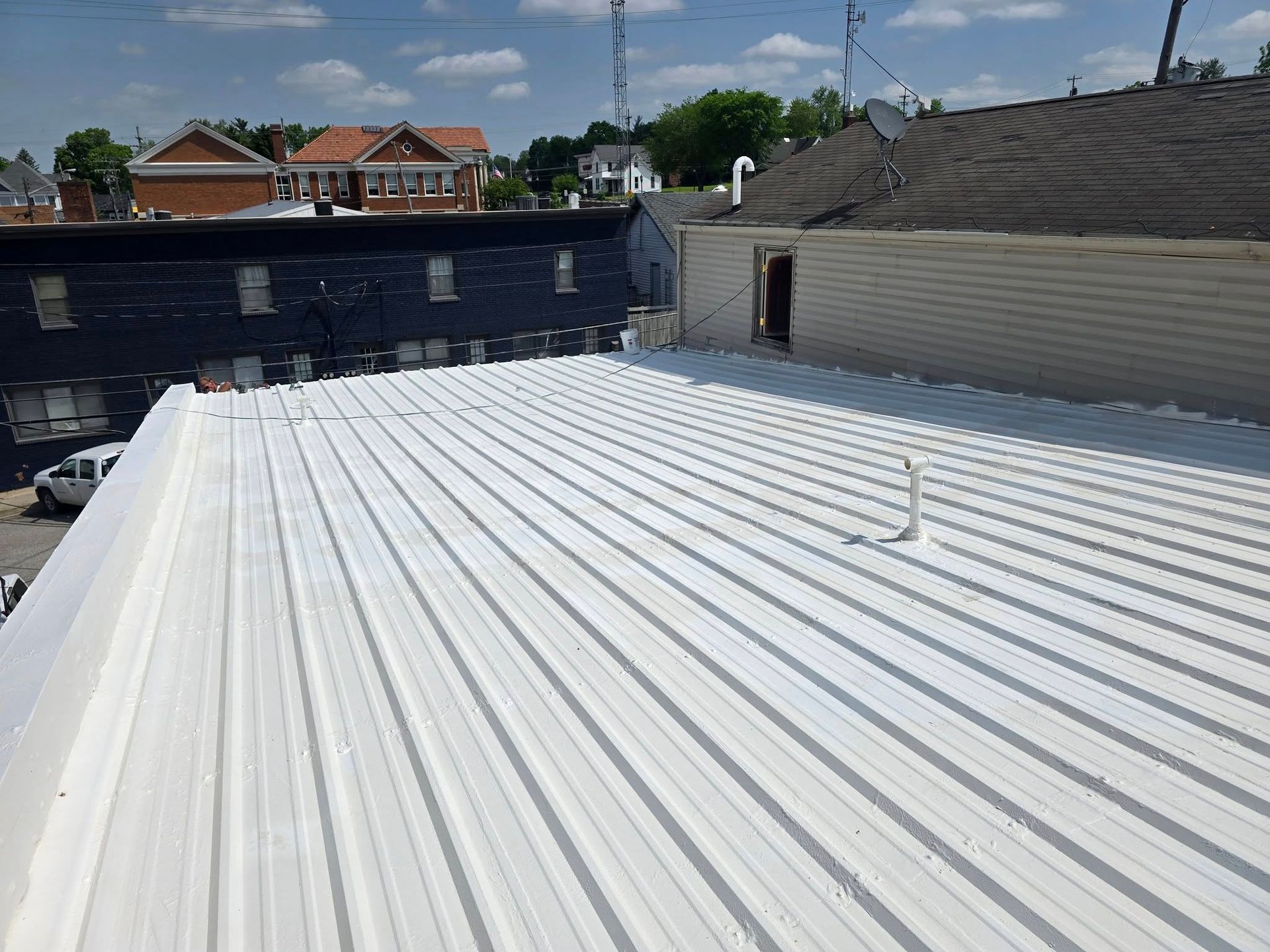 A bright white metal roof on a building, seen from a high vantage point, with neighboring rooftops and trees in the back.