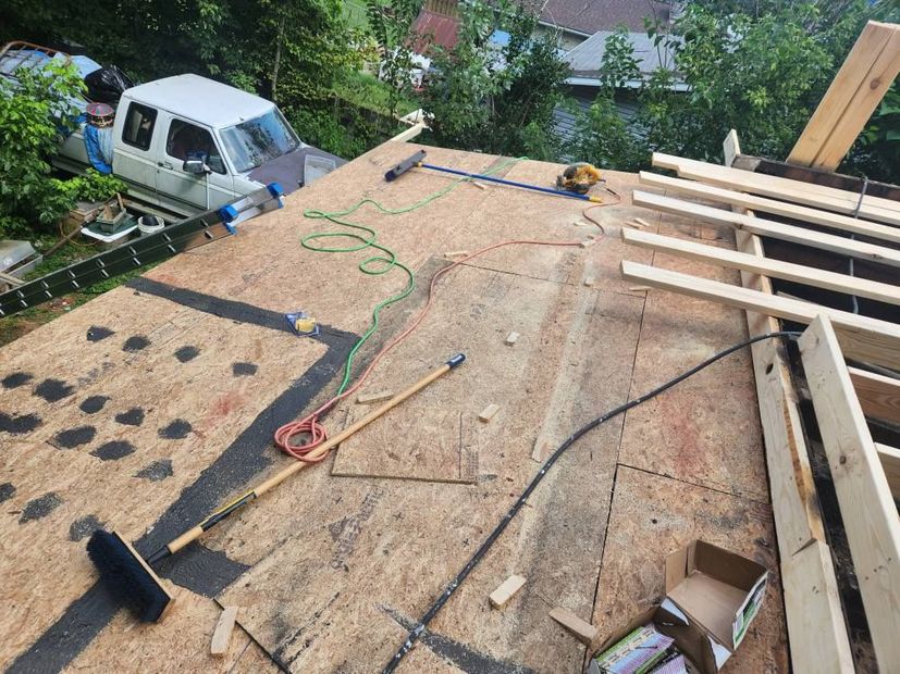 An elevated view of a plywood roof under construction, featuring scattered tools, building materials, and a parked truck.