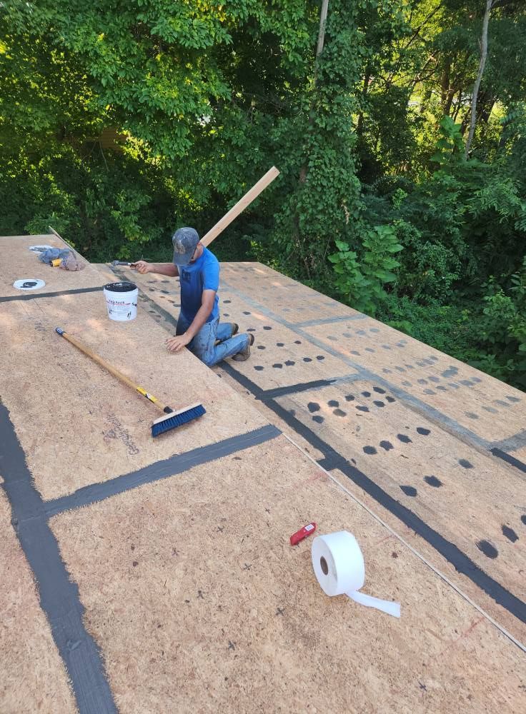 A worker in a blue shirt kneels on a plywood roof, applying black seam tape with tools nearby, surrounded by forest.