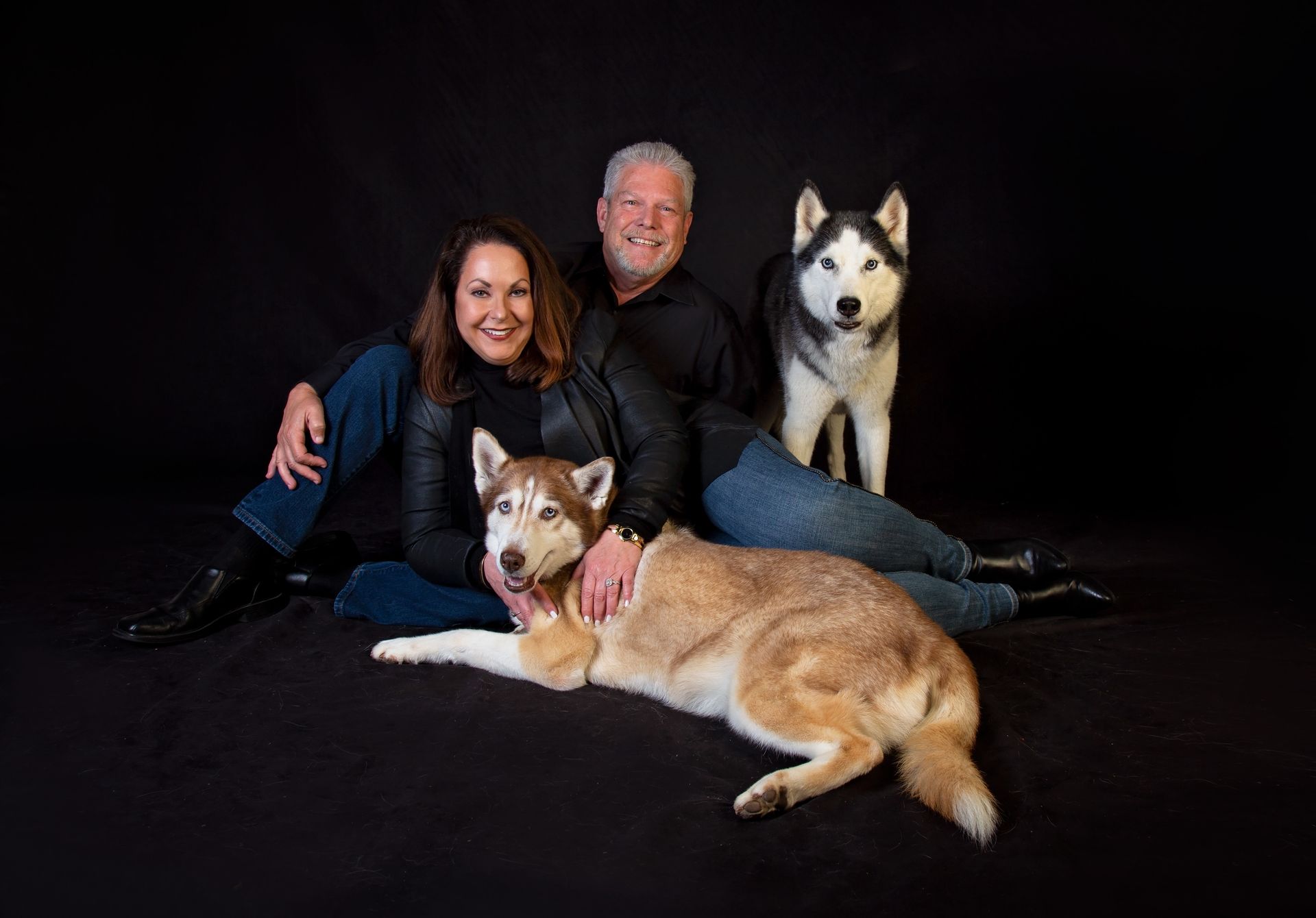 A man and a woman are posing for a picture with their two husky dogs.