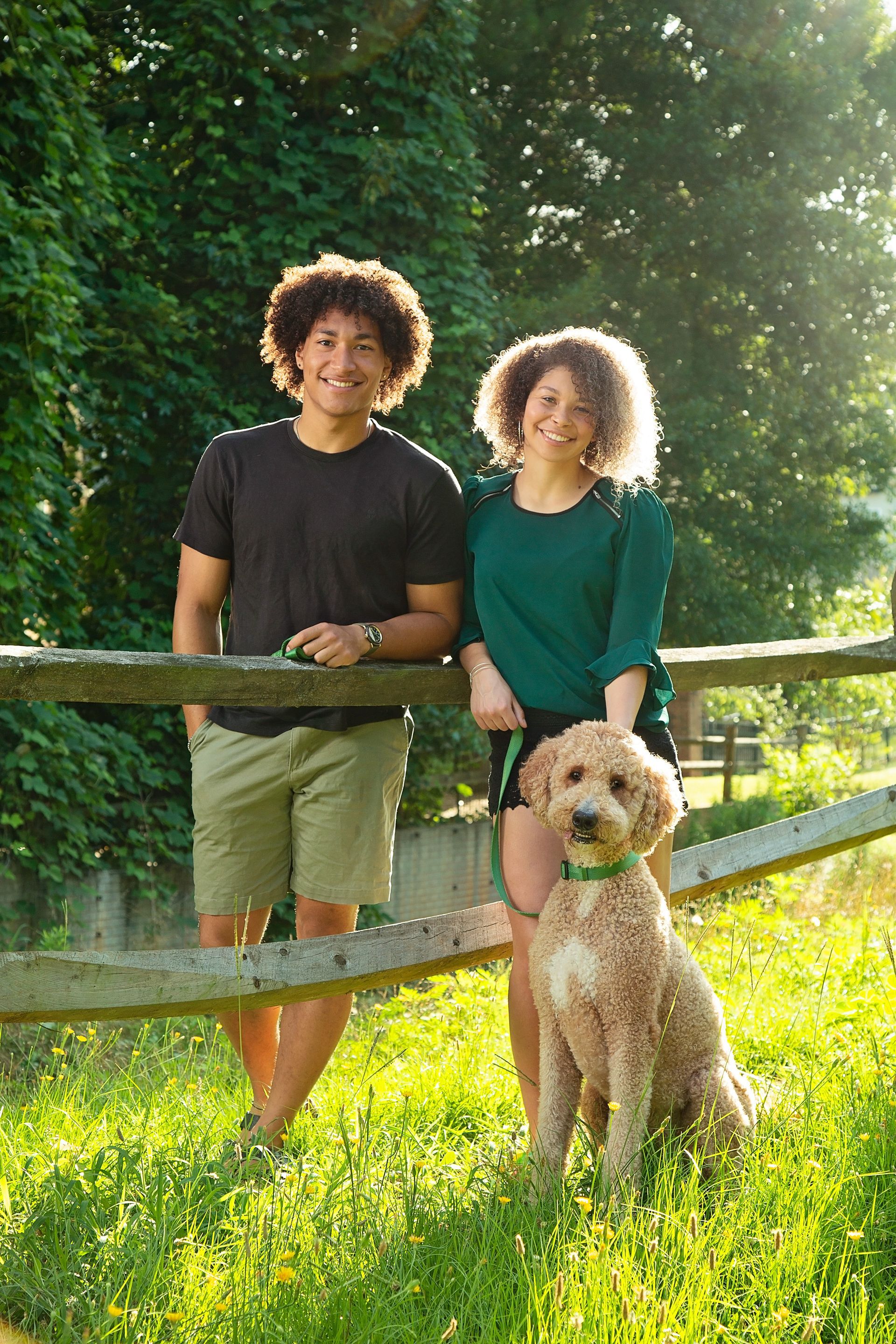 A teenage brother and sister are standing next to a wooden fence with their family dog.
