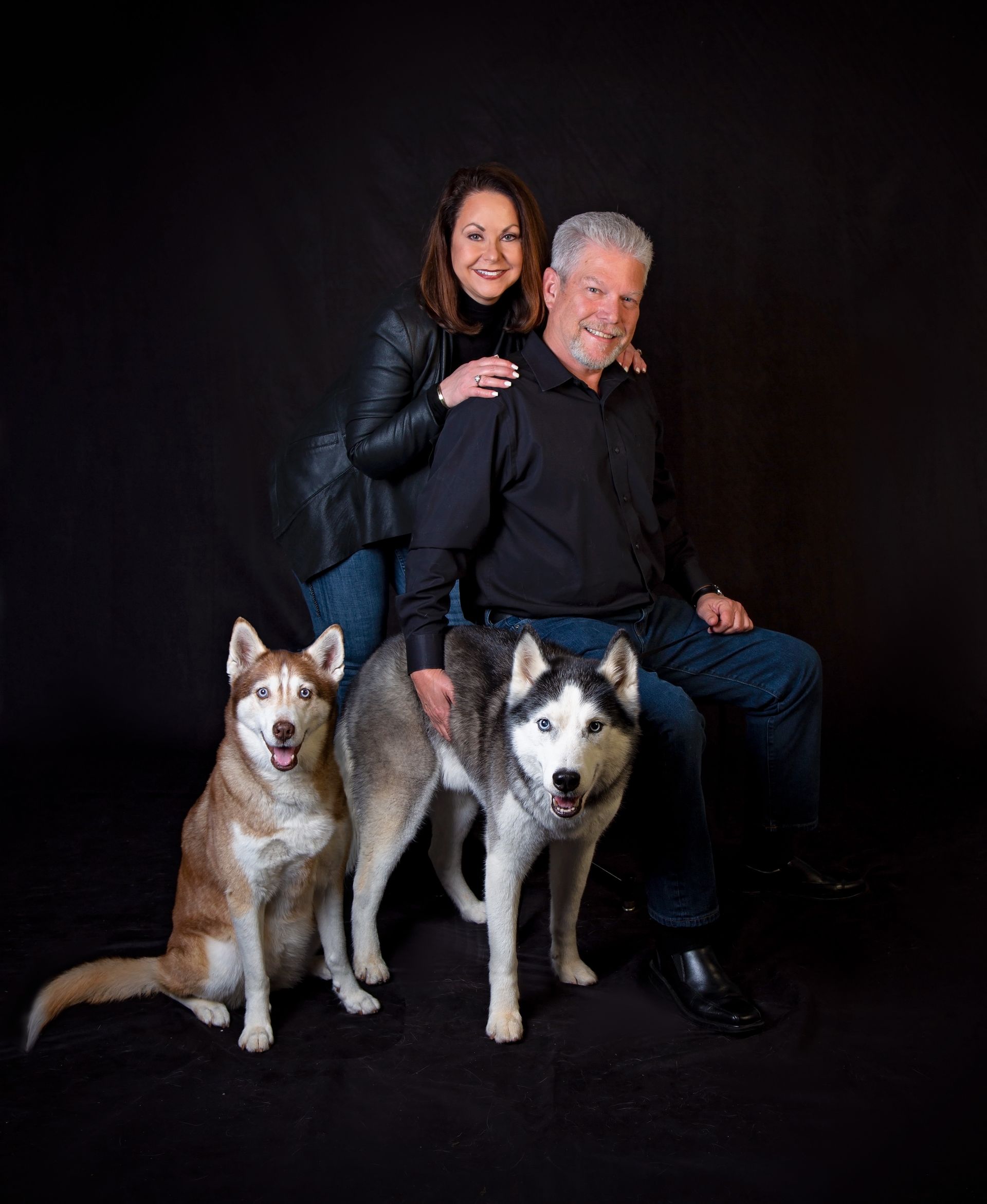 A man and a woman are posing for a portrait with two husky dogs.