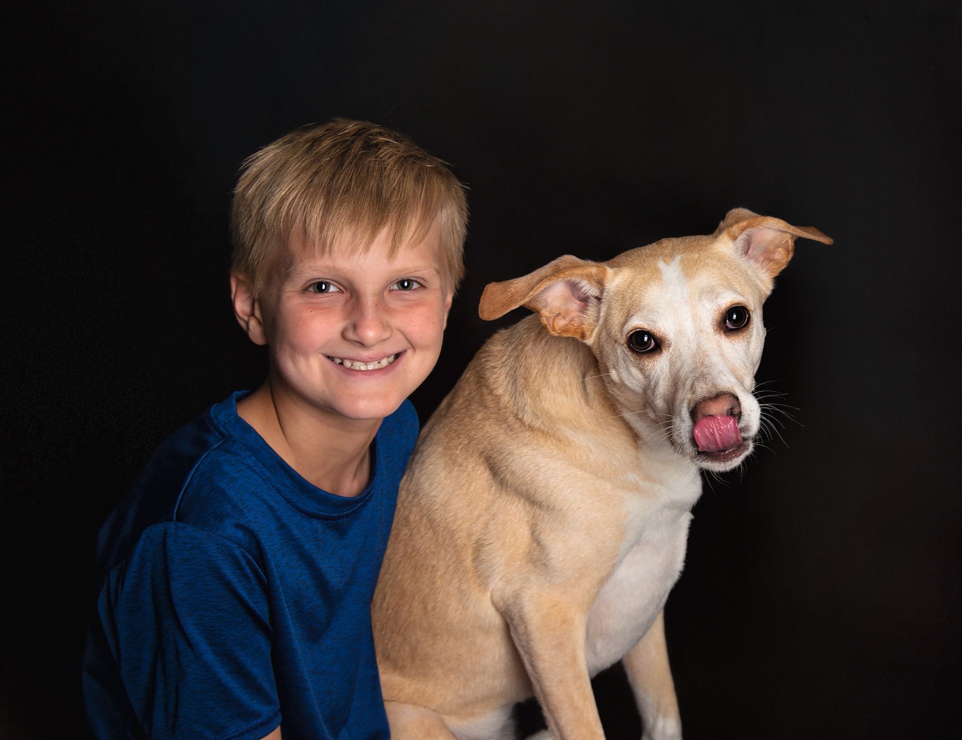A young boy is sitting next to a dog that is sticking its tongue out.