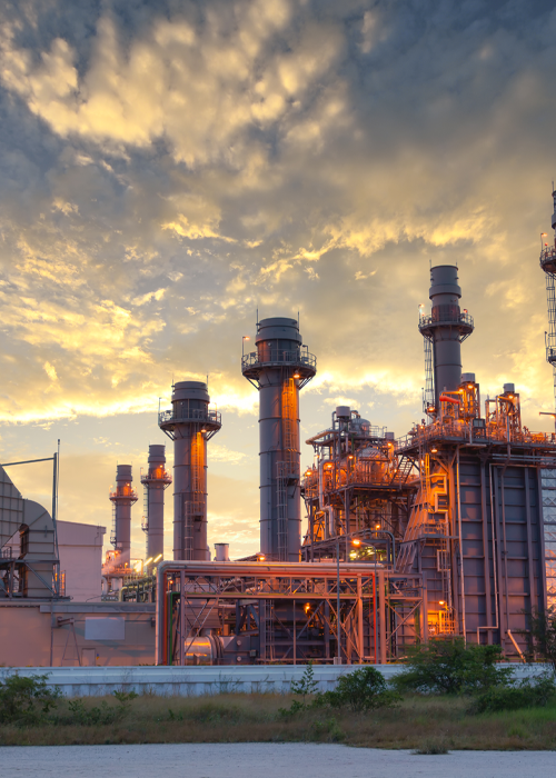Power plant at dusk, tall smokestacks and industrial structures against an orange and yellow sky.