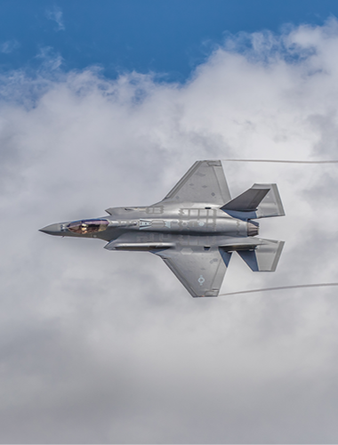 F-35 fighter jet in flight, gray against a cloudy blue sky, contrails visible.