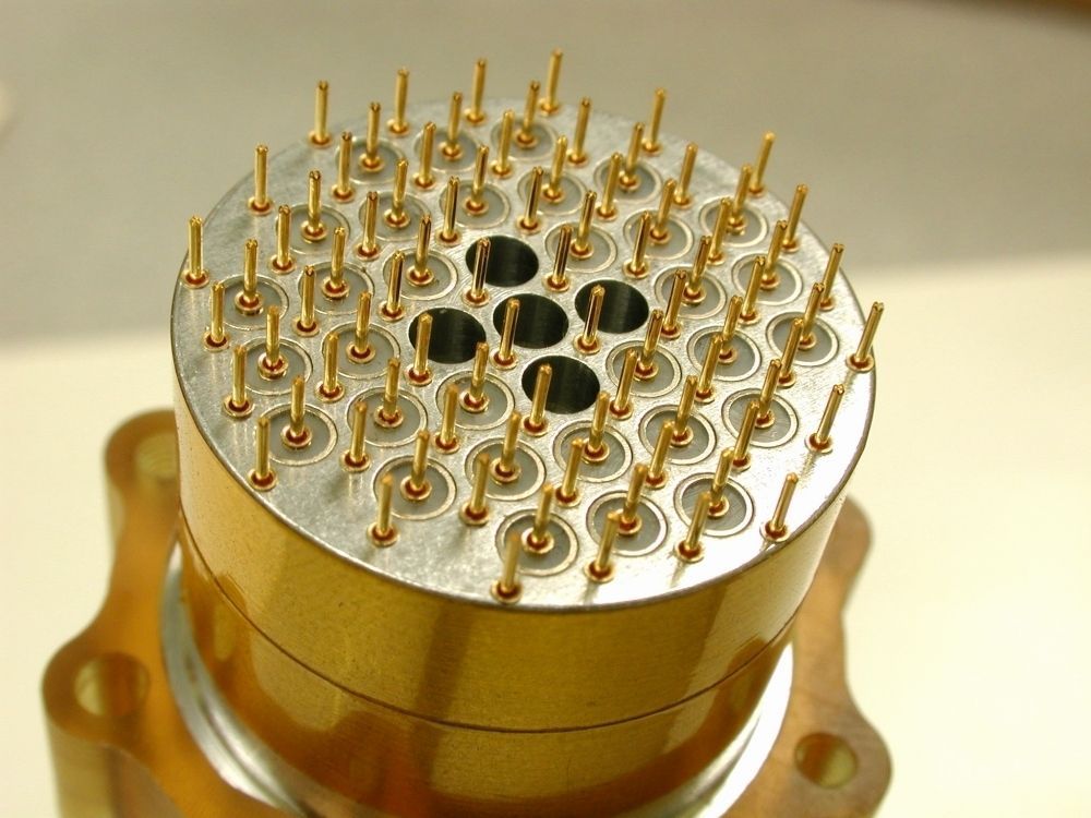 Close-up of a gold circular connector with numerous pins and several central holes.