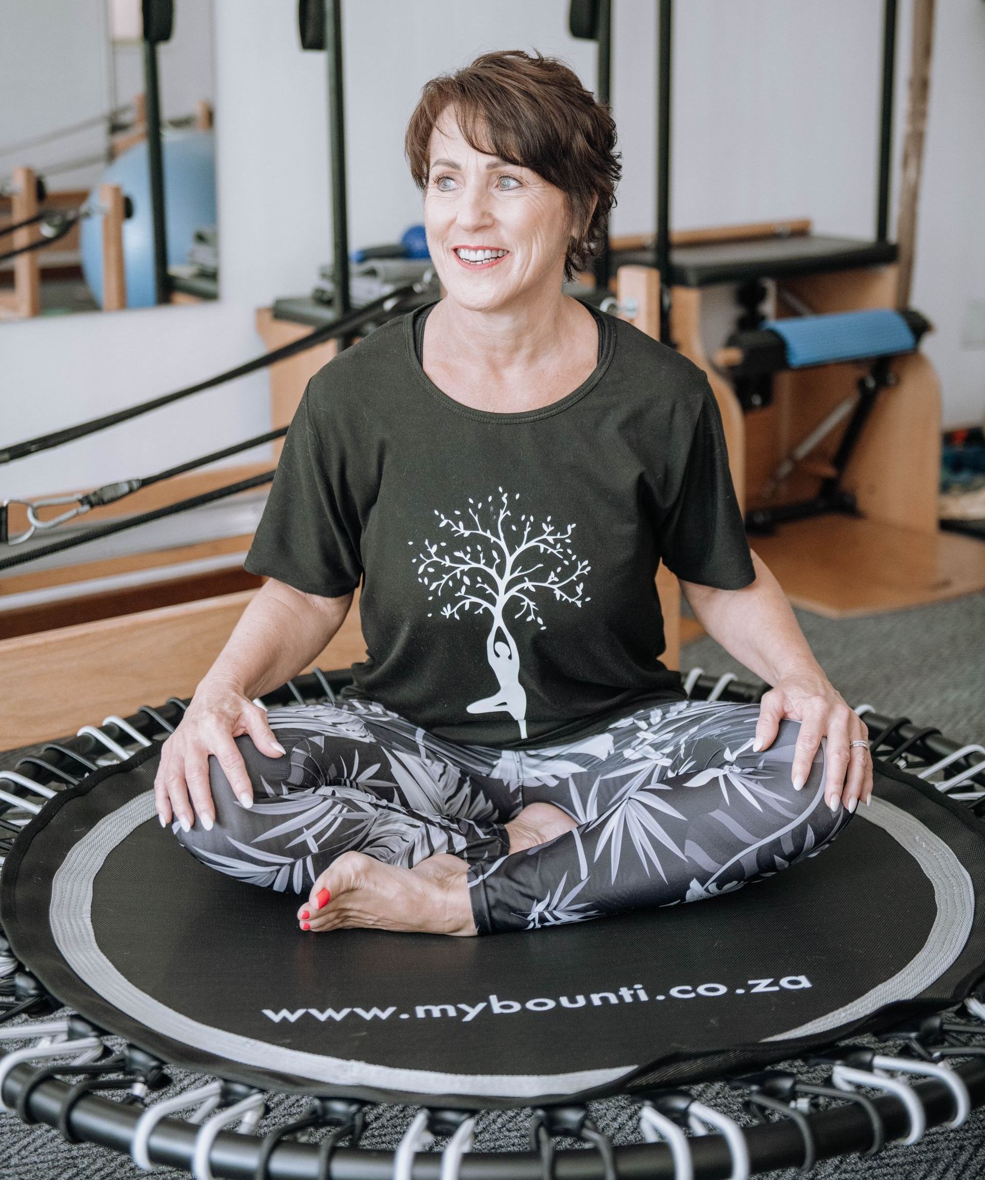 Woman seated cross-legged on a rebounder in a fitness studio, smiling and wearing a dark tree-print shirt.