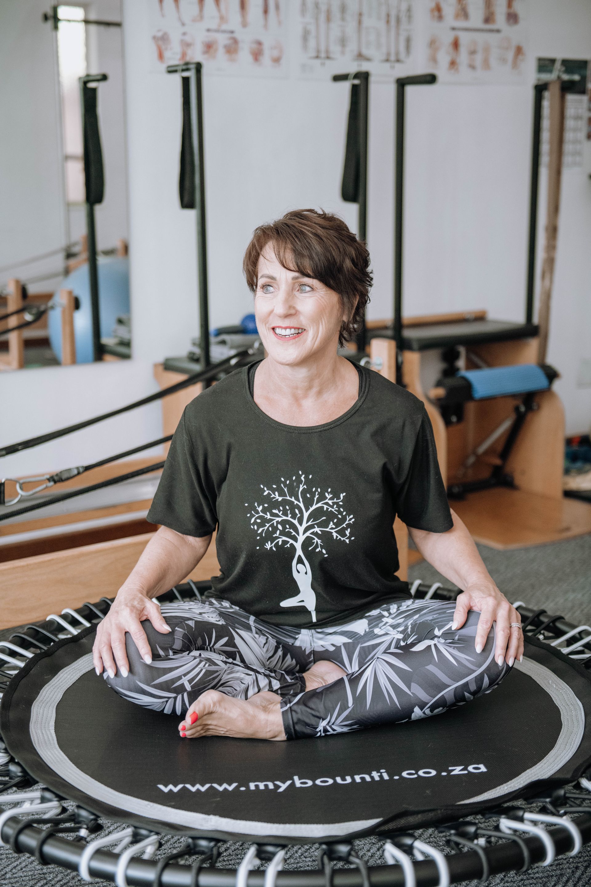 Person sitting cross-legged on a Pilates reformer mat in a studio, smiling.