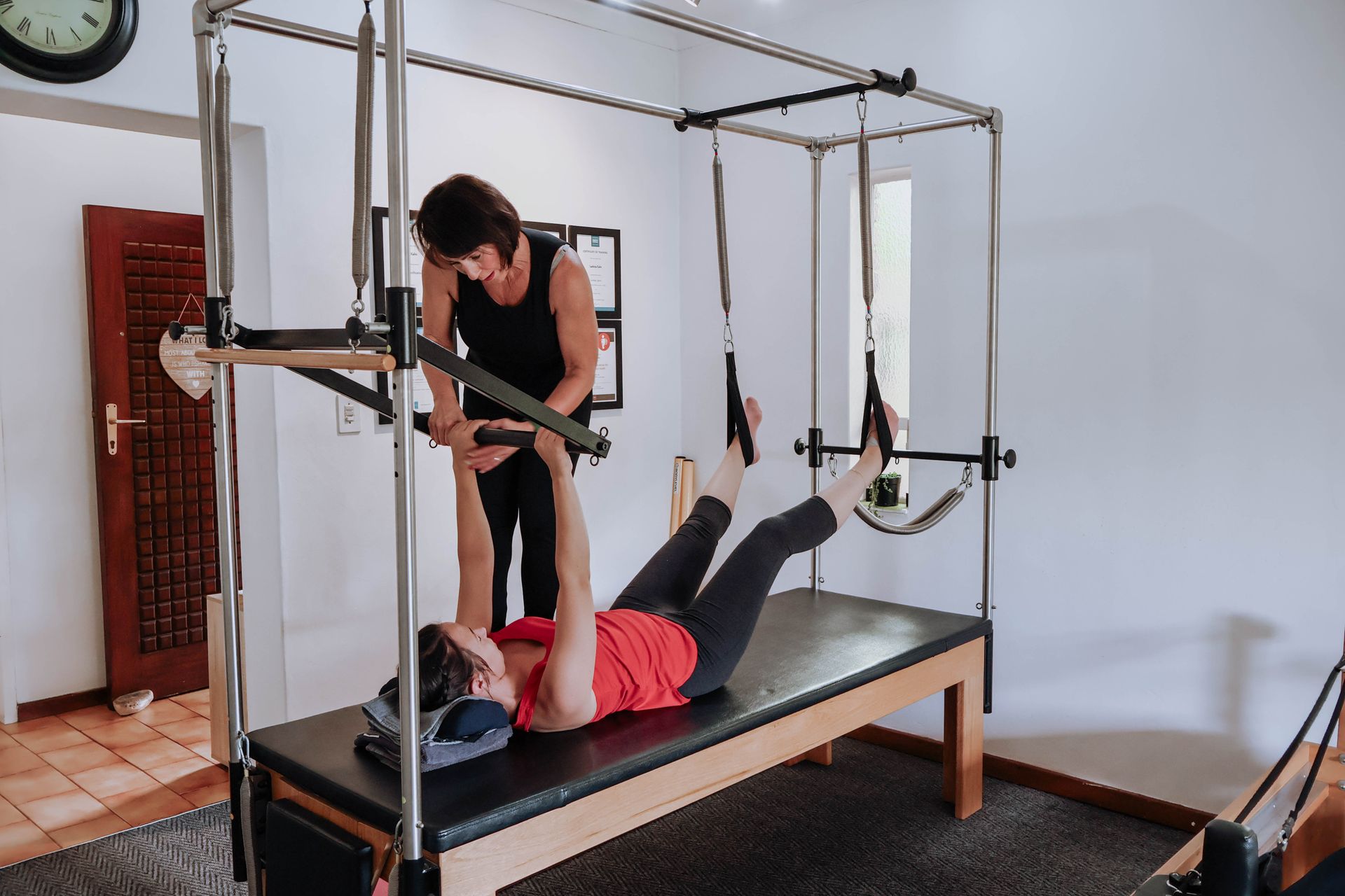 Person on a Pilates reformer adjusting a woman doing leg exercises in a bright studio