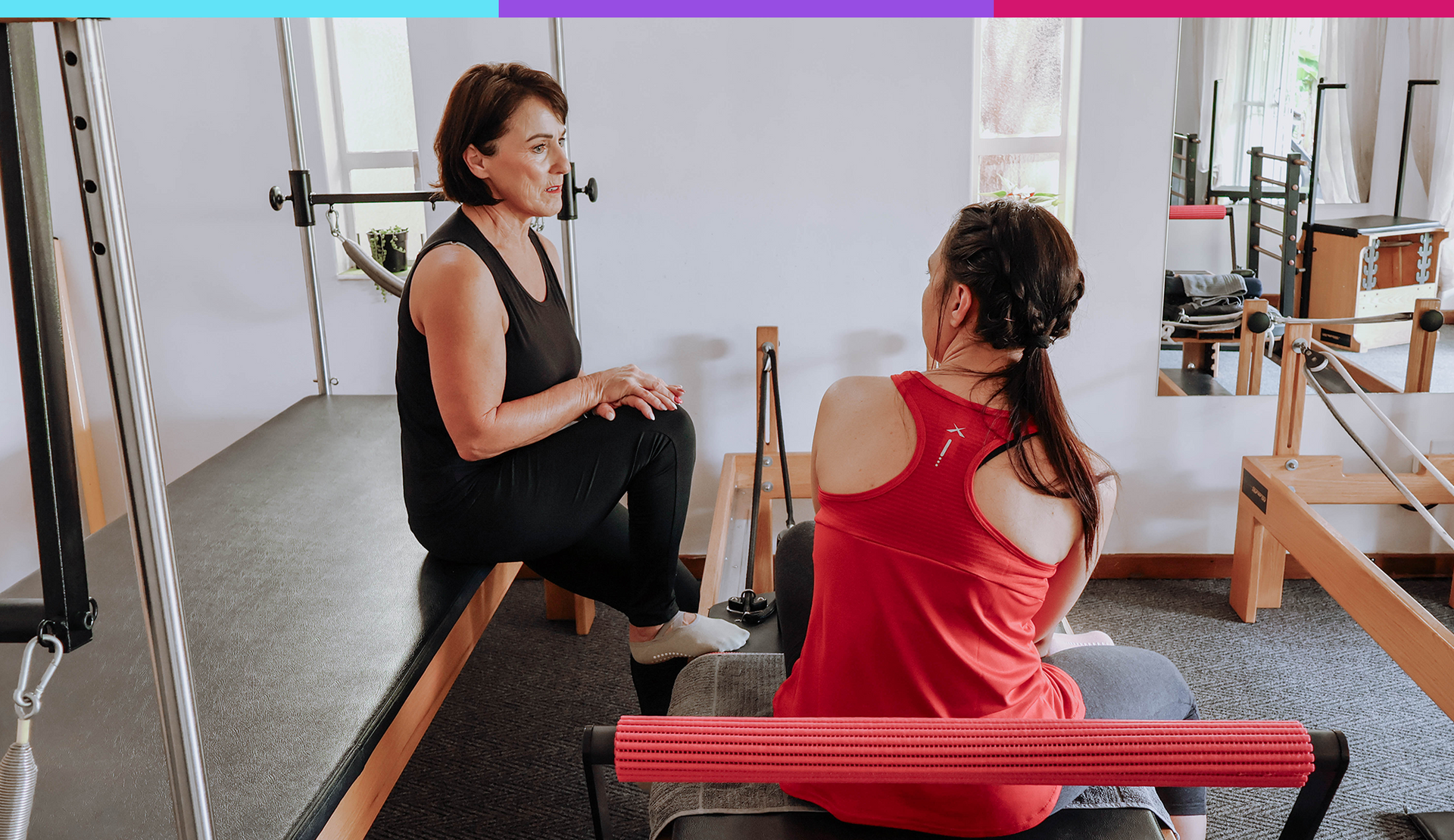 Two people talking on a Pilates reformer in a bright studio, one in black and one in red.