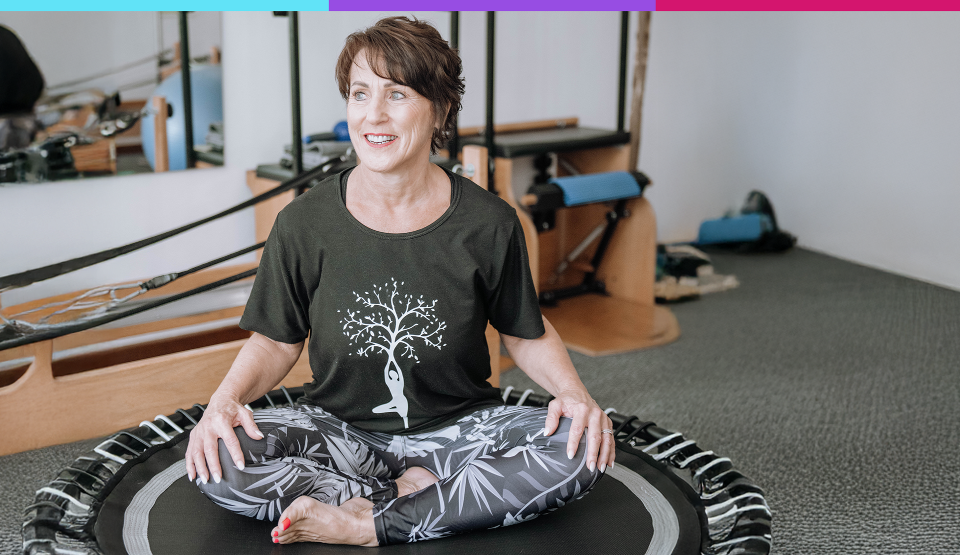 Person sitting cross-legged on a rebounder in a studio, smiling, with fitness equipment in the background