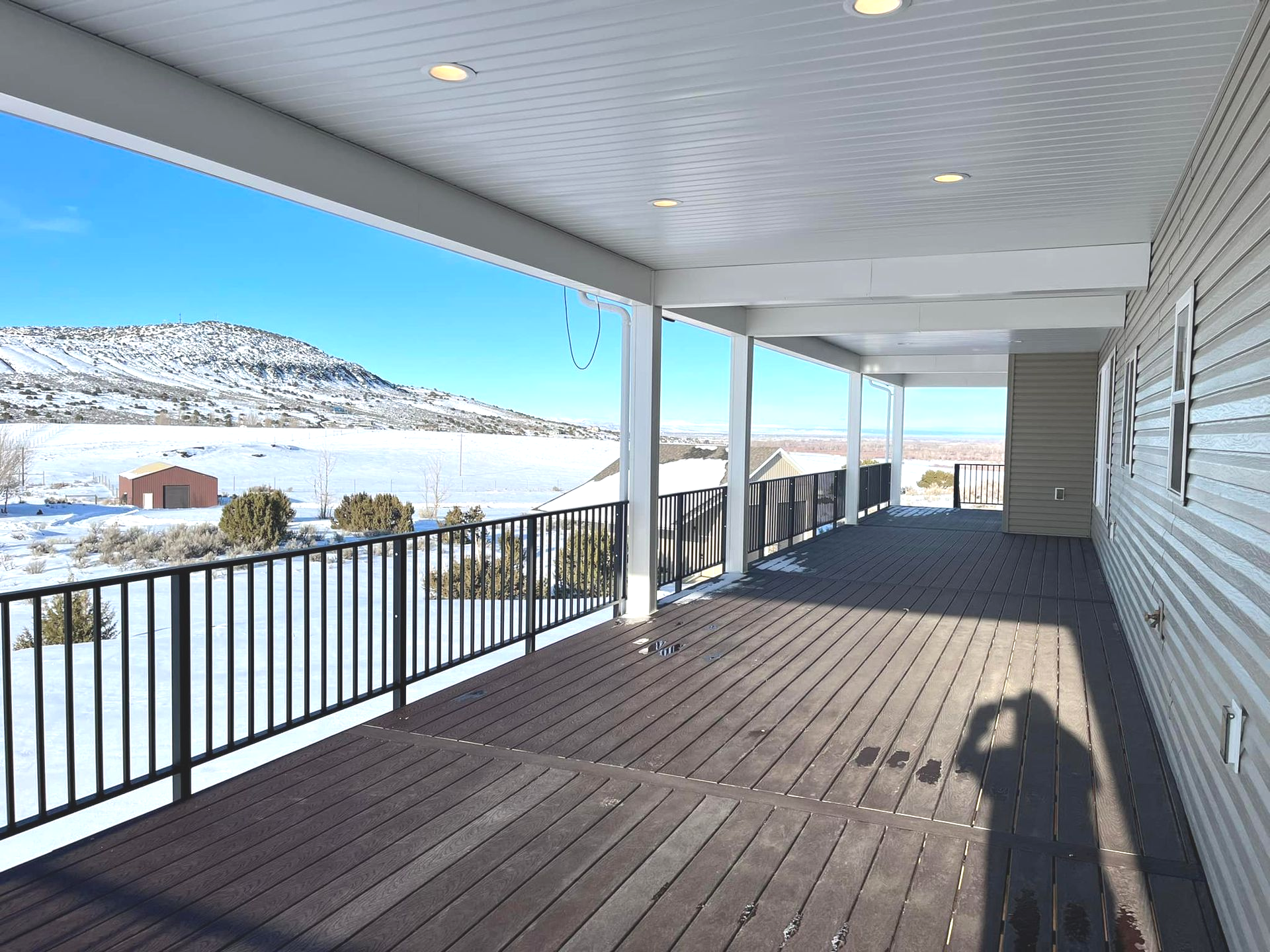 A covered wooden deck with a snowy mountain view. Black railing, light-colored siding, and bright blue sky.