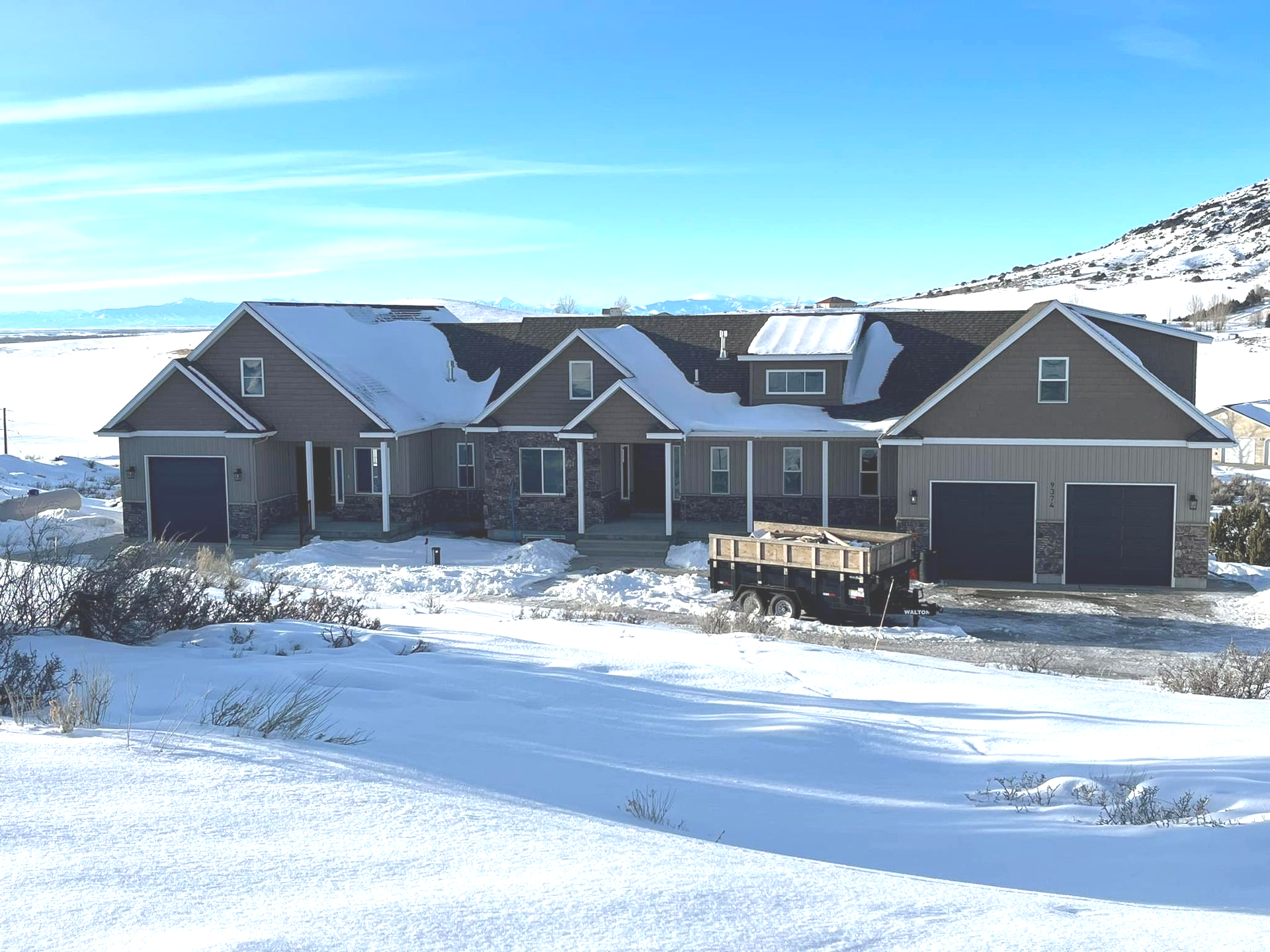 House with snow-covered roof and yard under blue sky, two-car garage, and mountain backdrop.
