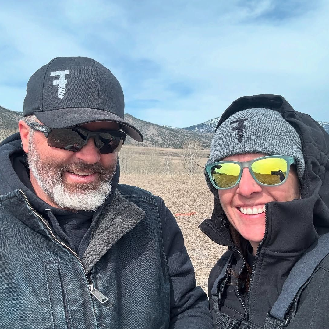 A smiling couple in winter attire outdoors, wearing hats with a logo. Mountains in the background.