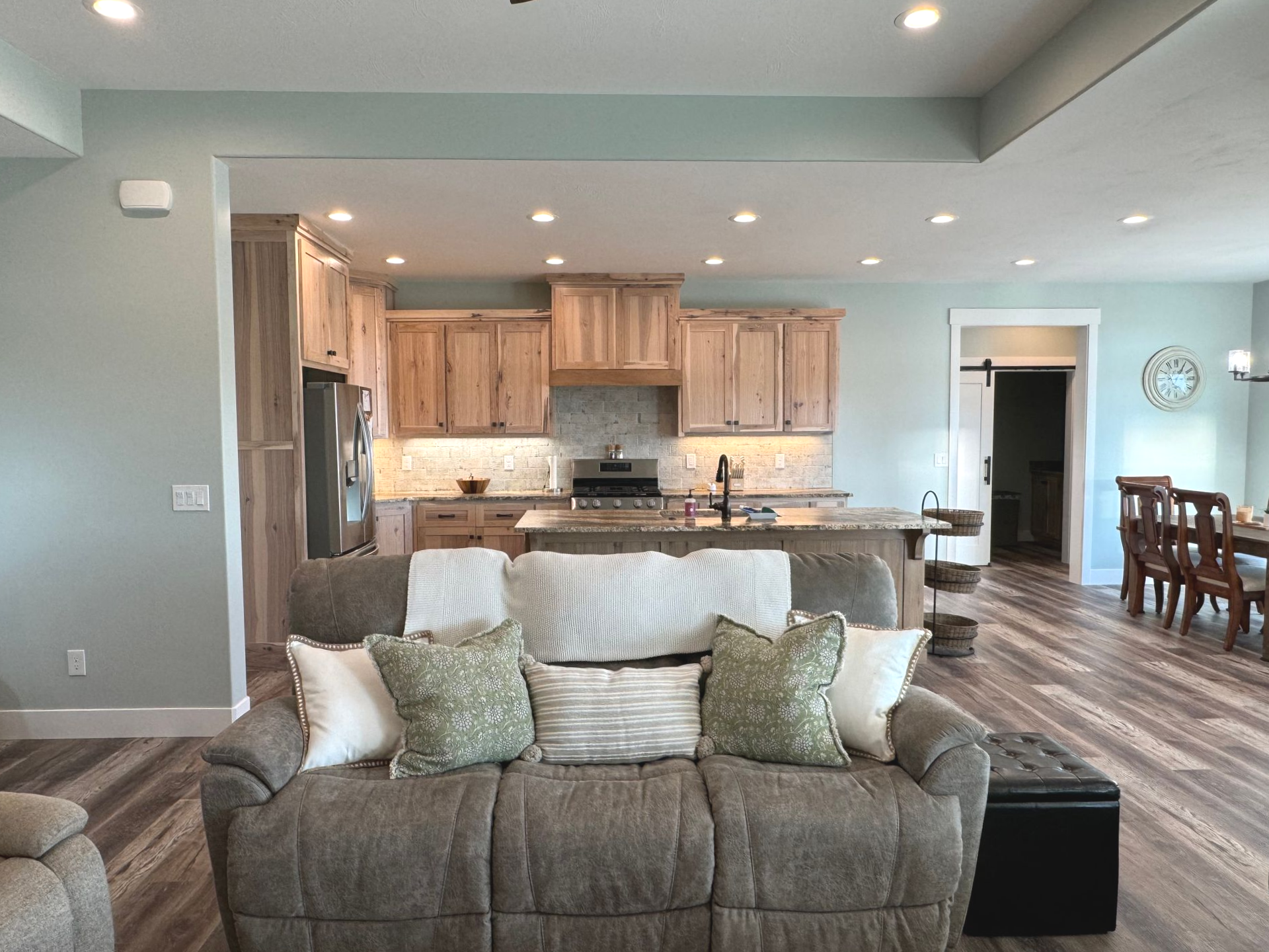Living room with kitchen in the background. Light wood cabinets, blue walls, a brown couch with pillows, and a dining area.