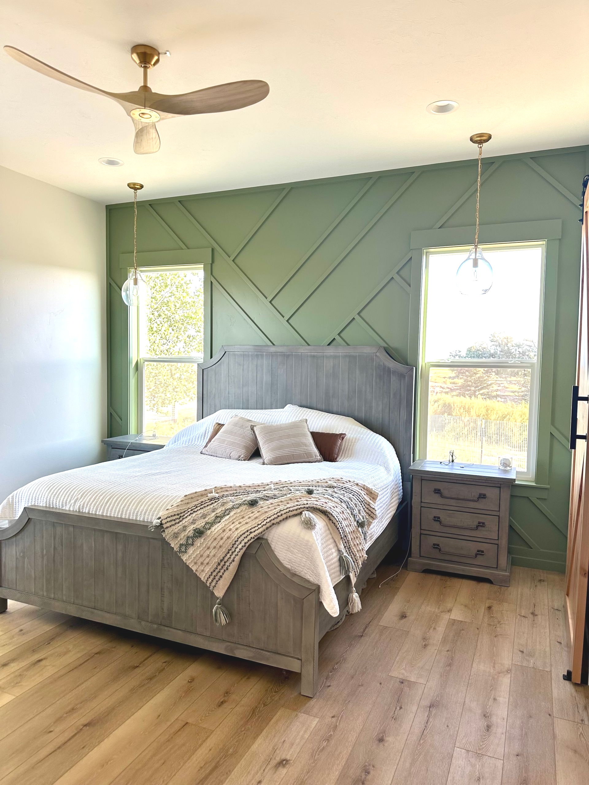 Bedroom with green accent wall, gray bed, light wood floors, two windows, and a ceiling fan.