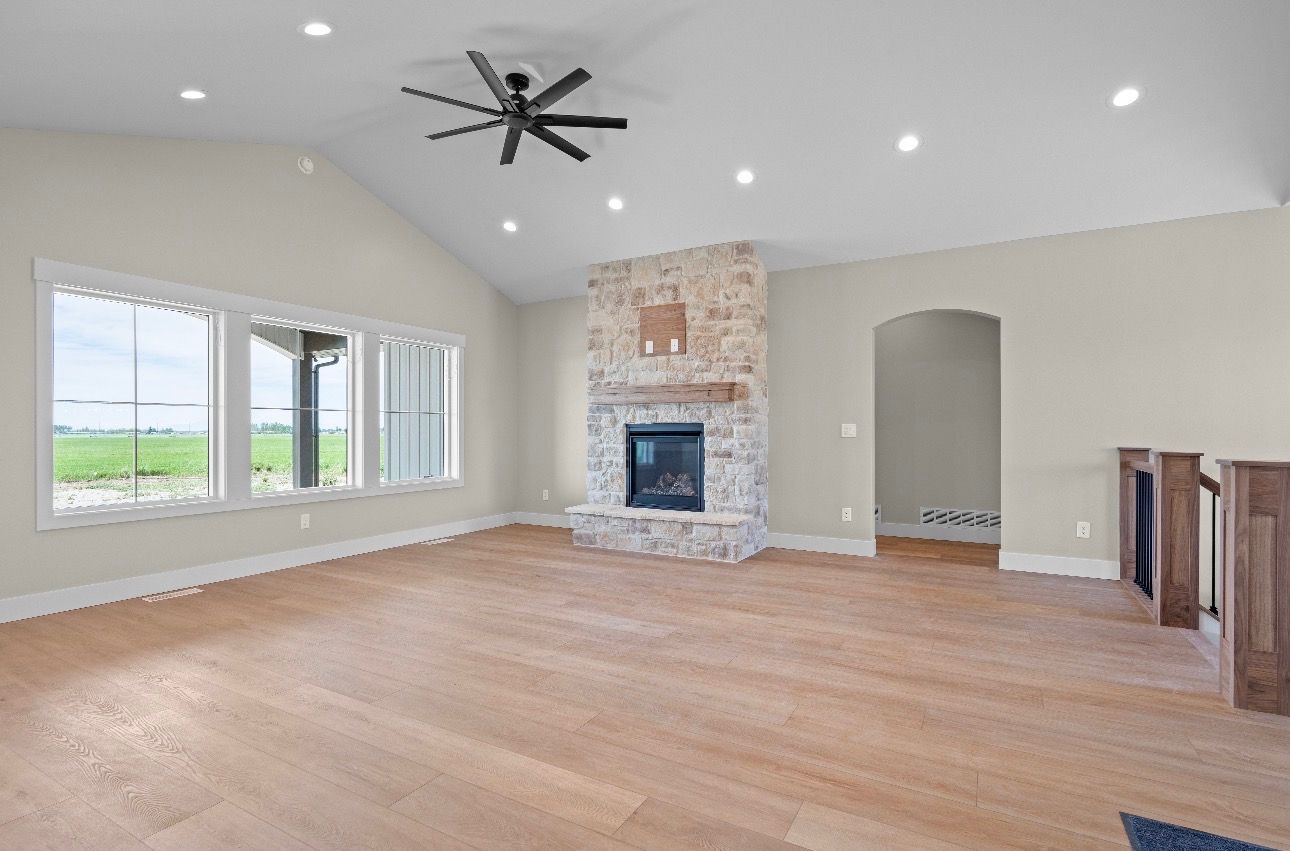 Spacious empty living room with vaulted ceiling, stone fireplace, large windows, and hardwood floors