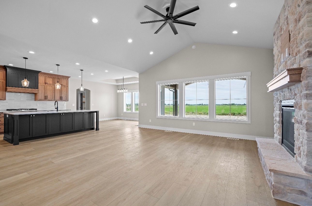 Spacious open-concept living room with kitchen, large windows, ceiling fan, and stone fireplace