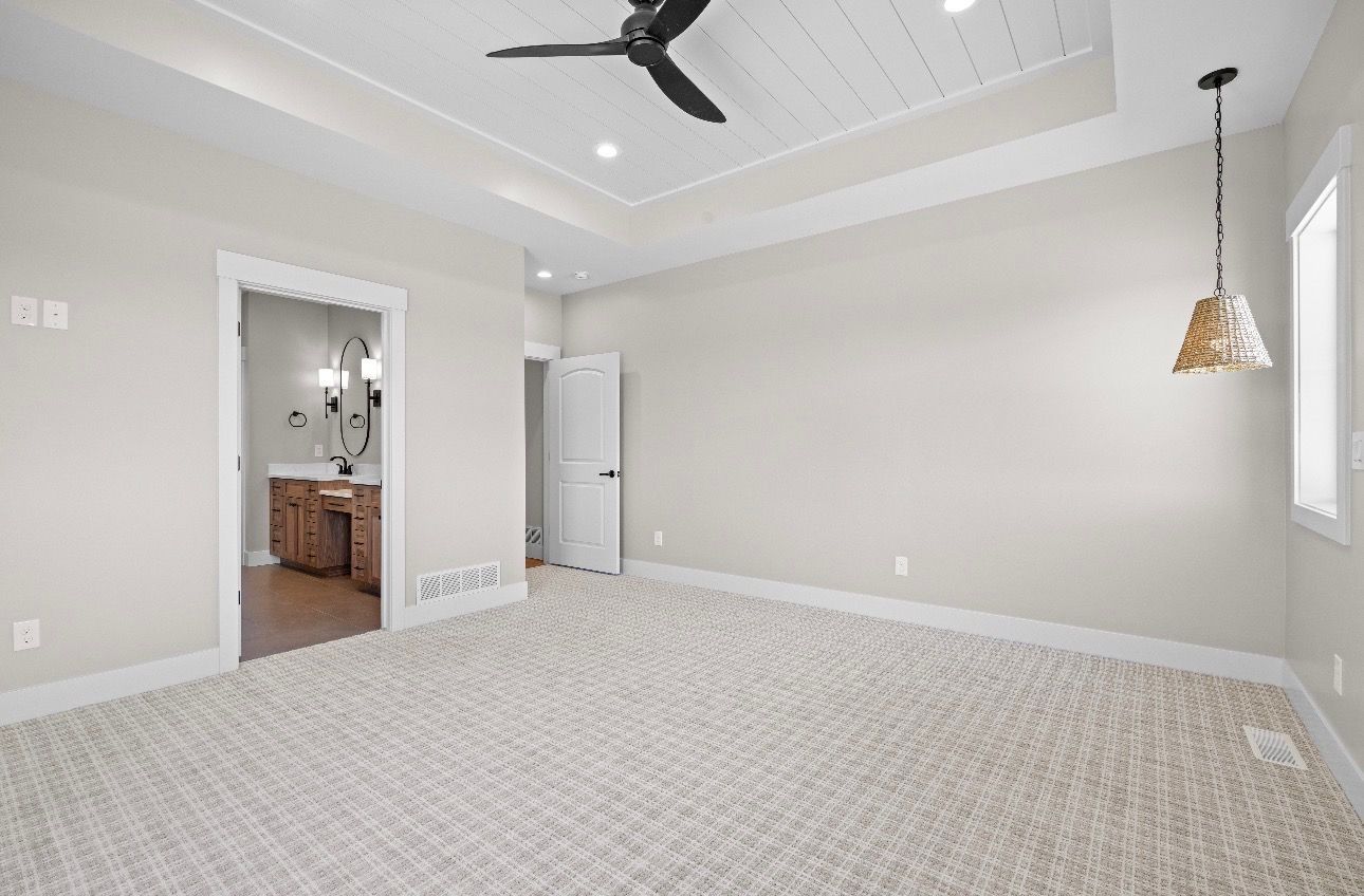 Empty beige bedroom with carpet, ceiling fan, pendant light, and doorway to a bathroom