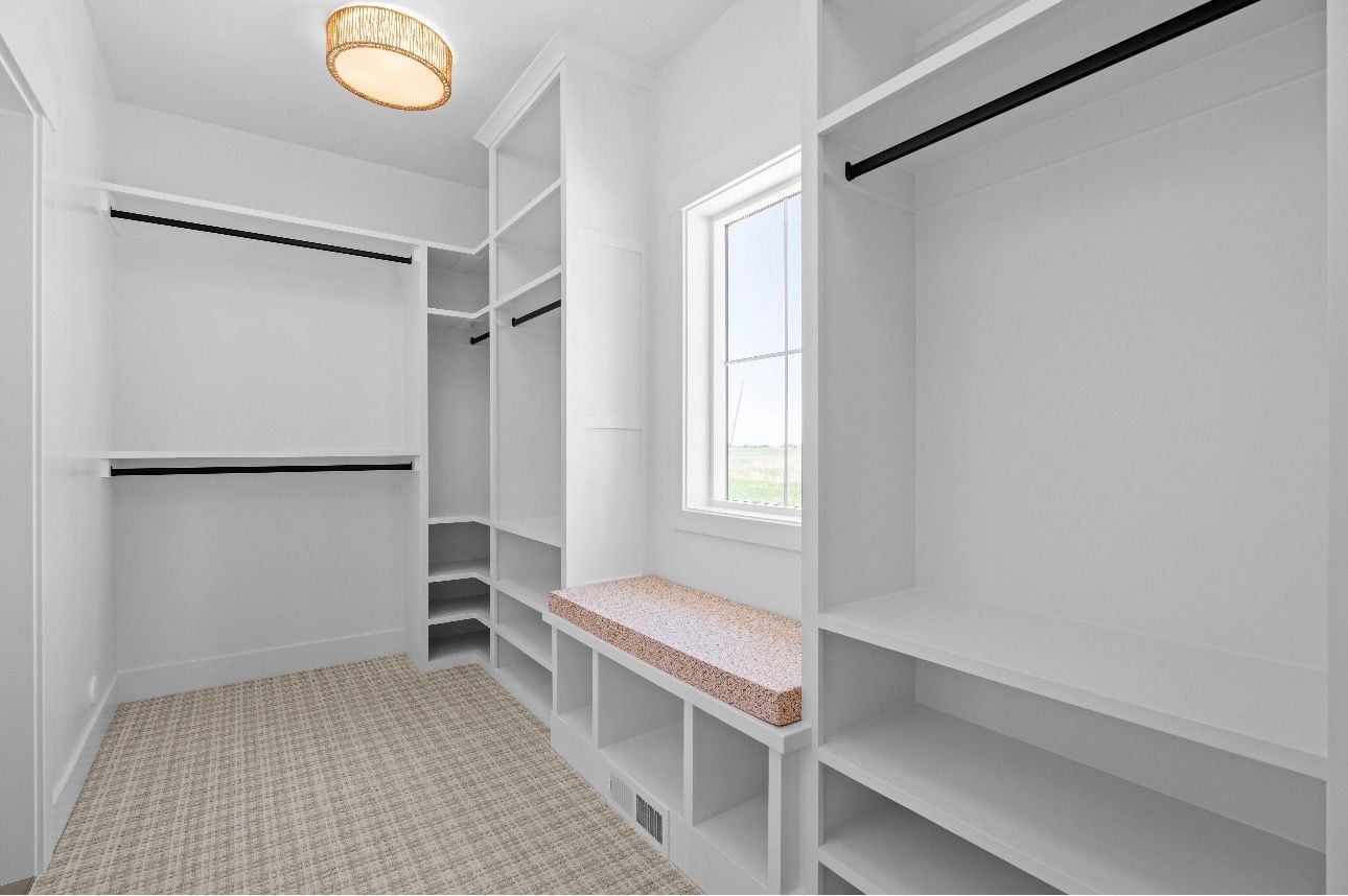 Bright walk-in closet with white built-in shelving, hanging rods, and a window seat by a window.
