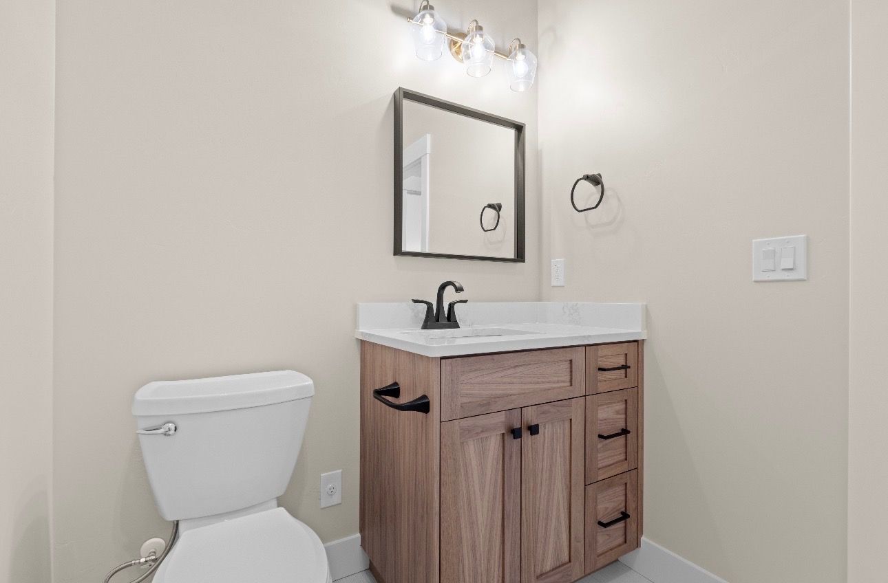 Small bathroom with toilet, wood vanity, sink, mirror, and wall light.