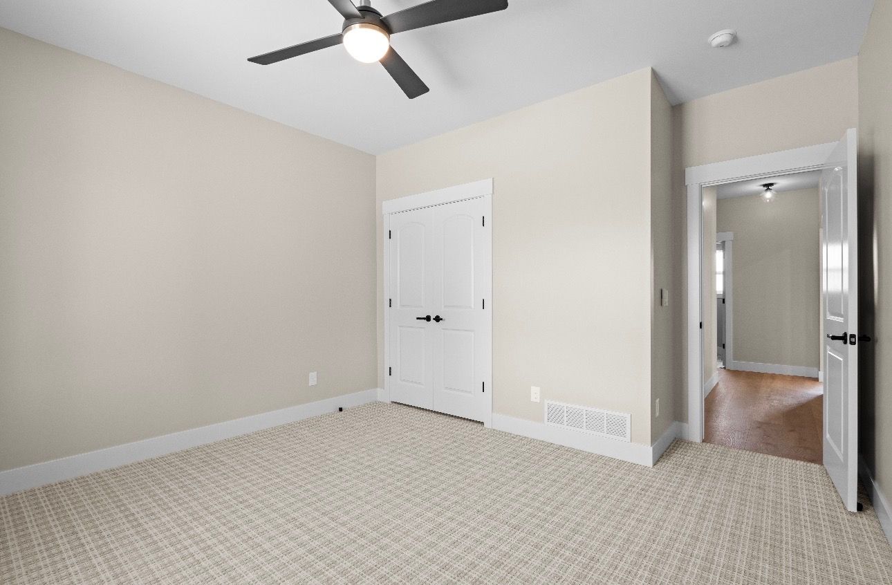 Empty beige bedroom with carpet, ceiling fan, closet doors, and an open doorway to a hall