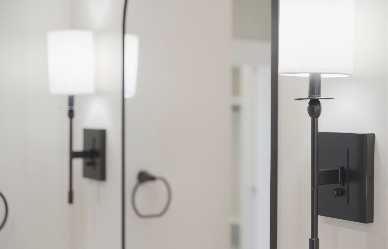 Wall sconce with white shade and black mount reflected in a mirror in a bathroom