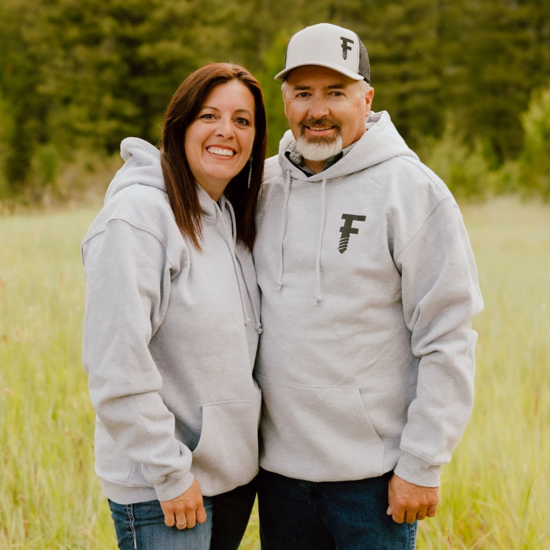 Couple in gray hoodies and a baseball cap, smiling in a grassy field.