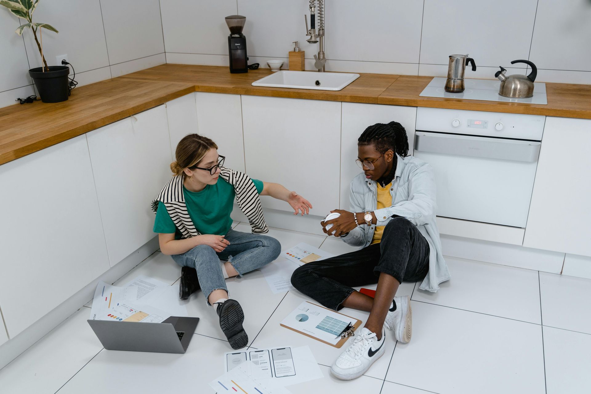 Two people sit on the kitchen floor, looking at papers and a laptop, planning for hardwood floor services in Port Washington